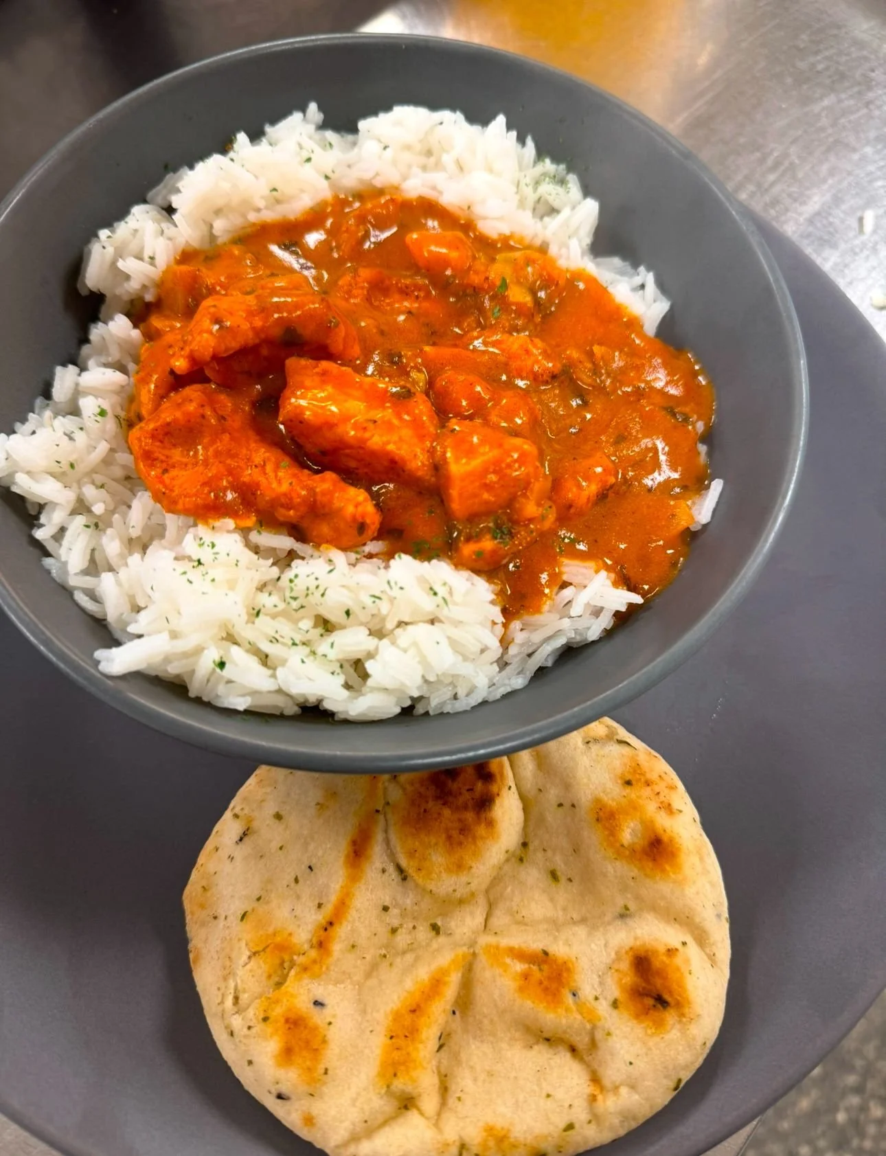 A bowl of white rice topped with chicken curry in sauce, with a piece of naan bread on a gray plate underneath.