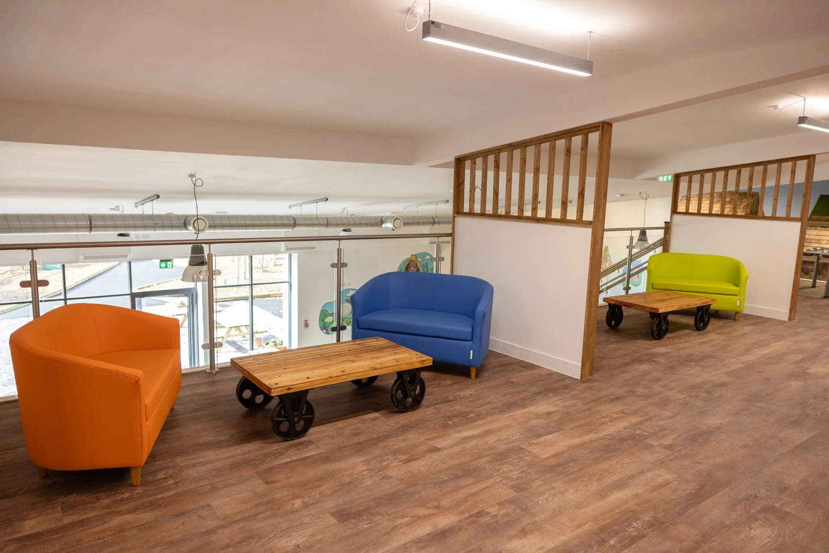 Colorful seating area with orange, blue, and green couches, wooden tables on wheels, inside a modern space with large windows, wooden flooring, and partial wooden walls.