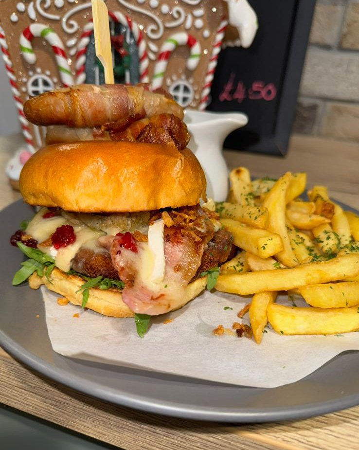 Close-up of a bacon cheeseburger with fries on a gray plate, with a gingerbread house decoration in the background.