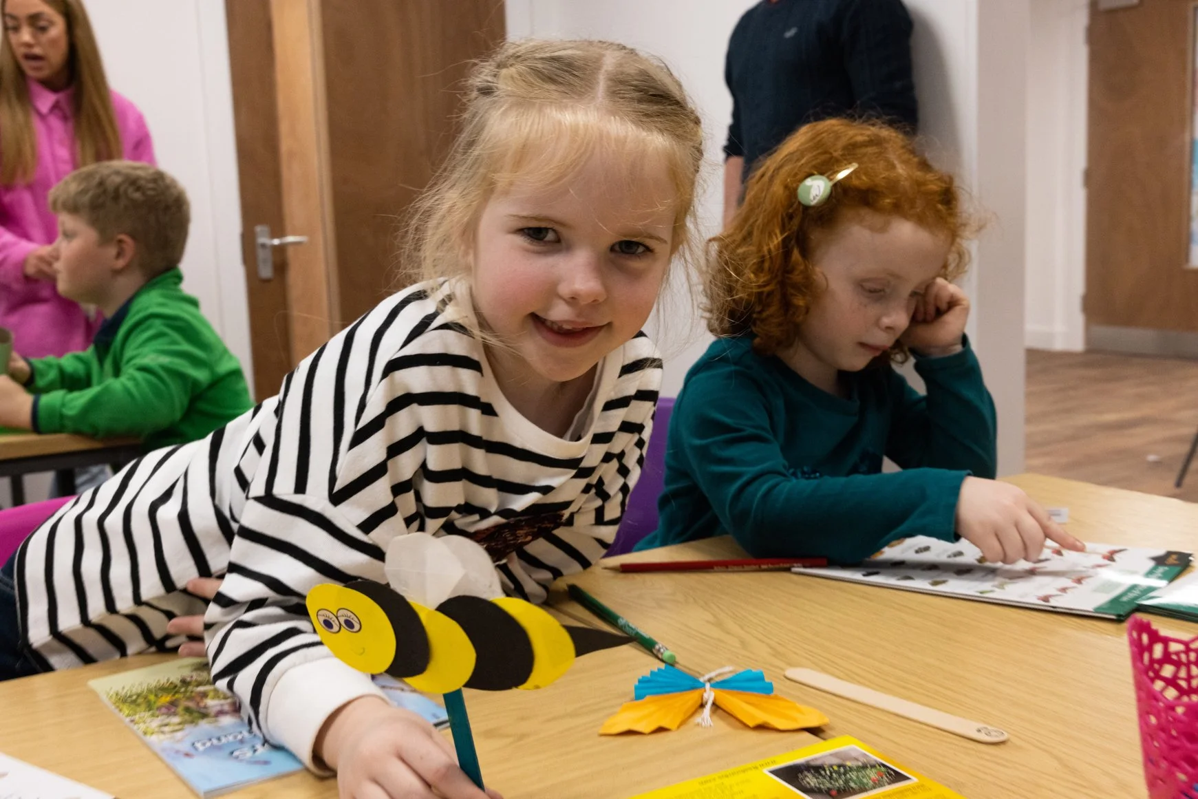 Two young girls at a table with educational and craft materials, one smiling at the camera and the other focused on a magazine, with colorful paper butterflies and a bee craft on the table.