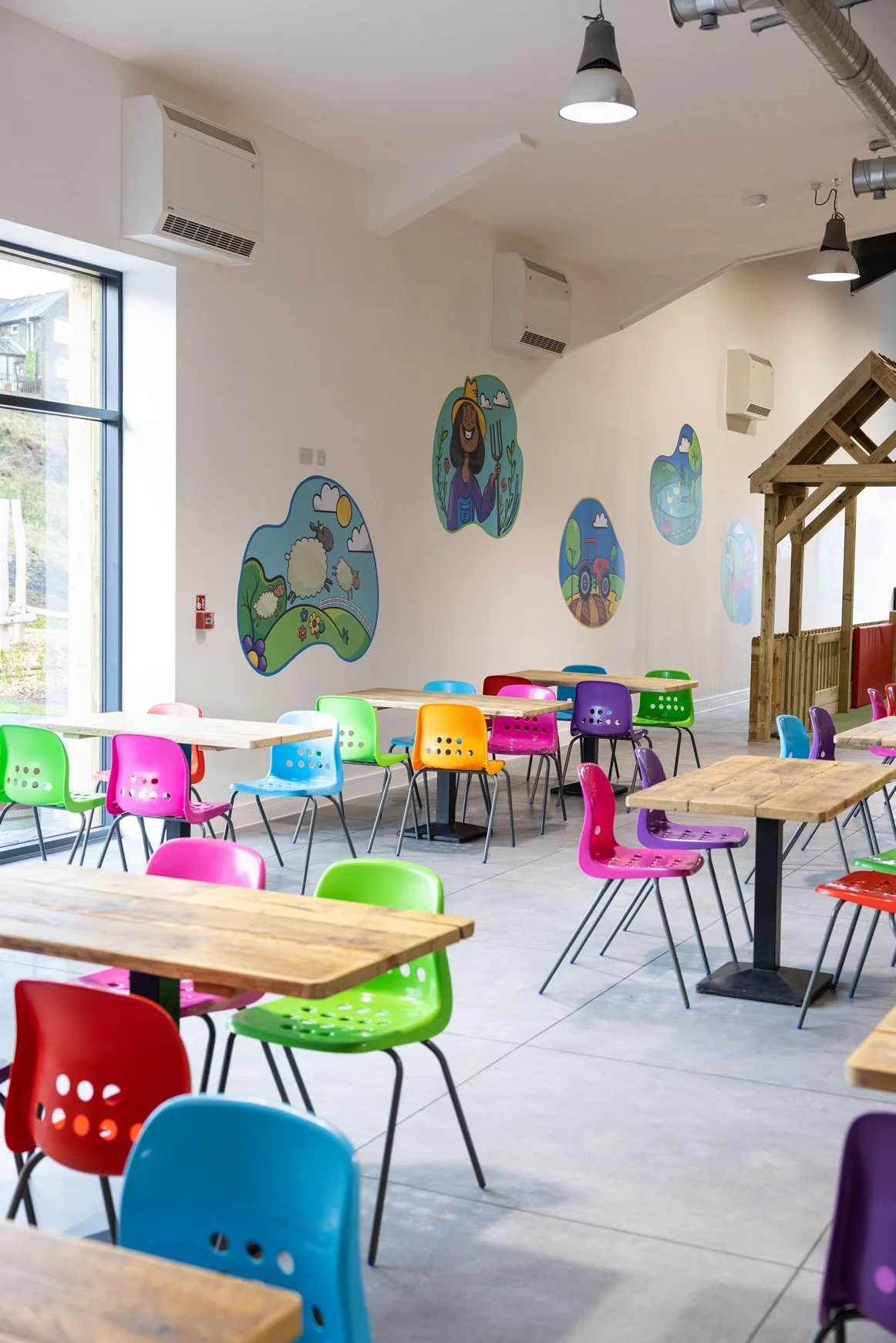 Brightly colored chairs in green, pink, blue, orange, purple, and red around wooden tables in a cheerful, modern indoor space with children's artwork on the white wall.