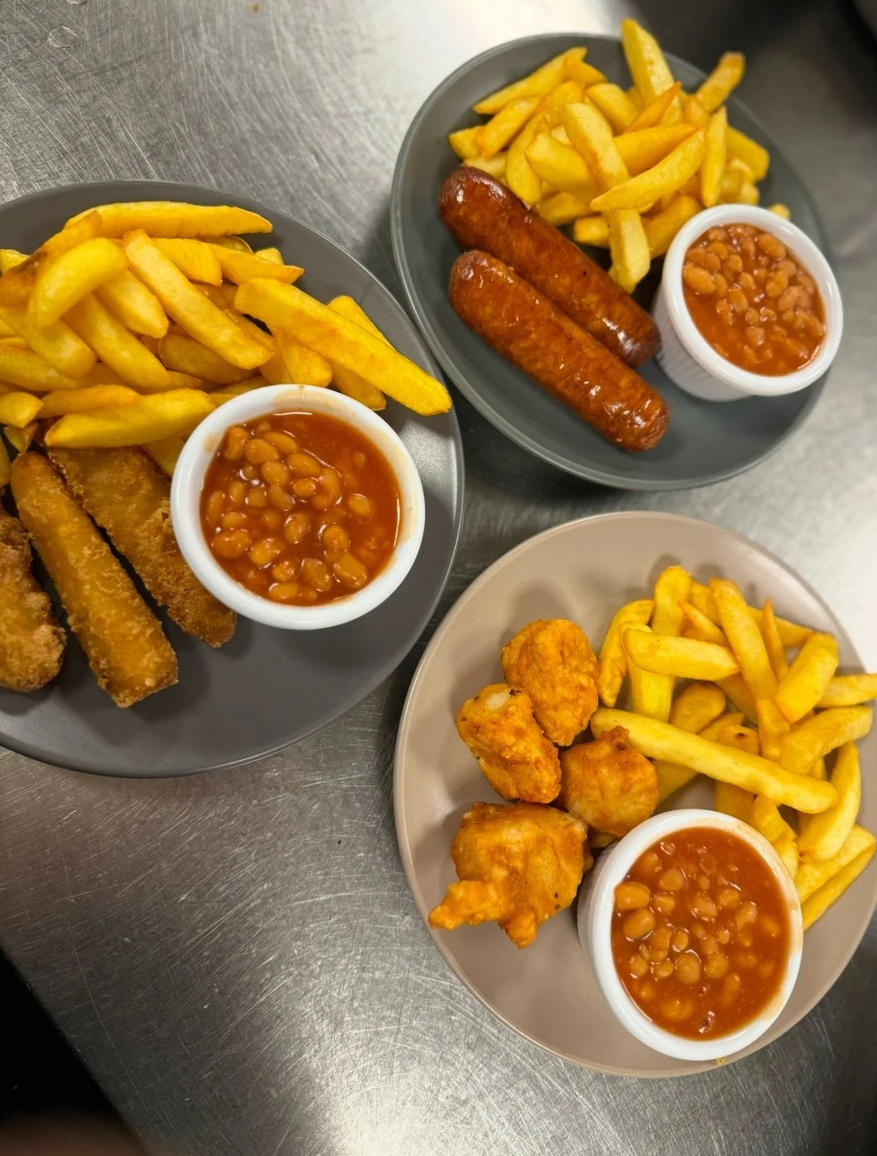 Three plates of fish and chips with French fries, baked beans, and sausages or chicken nuggets on a stainless steel table.