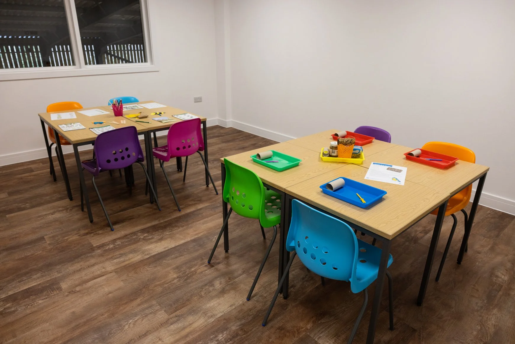 Two tables with colorful chairs and school supplies, including trays, paper, and art materials in a classroom setting with wooden flooring and white walls.