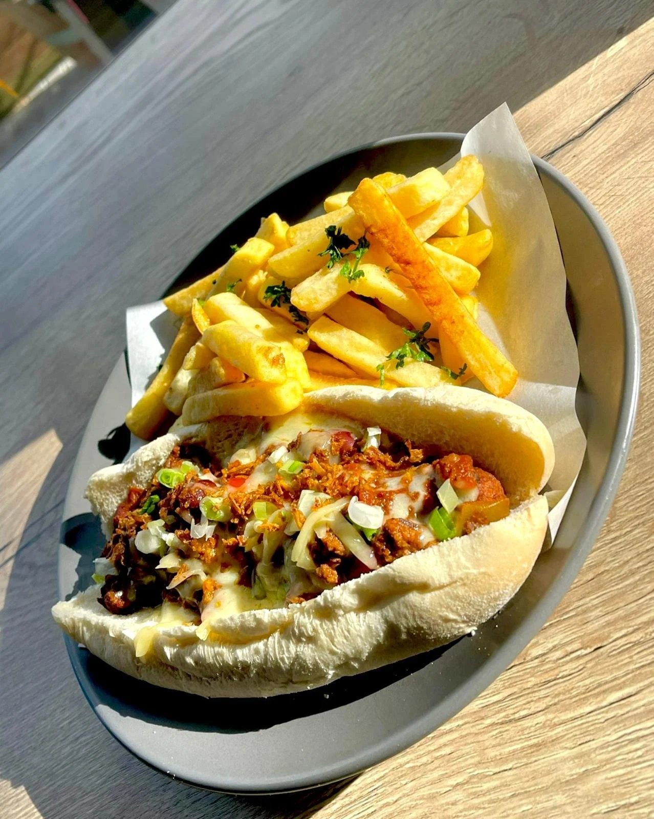 French fries and a sandwich with chopped herbs and shredded cheese in a steamed bun, served on a black tray lined with white parchment paper, on a wooden table.