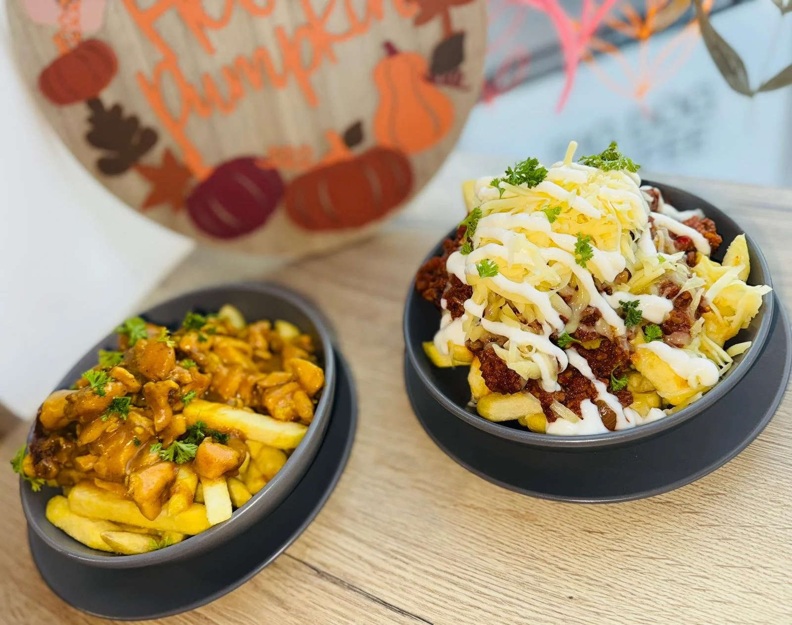Two bowls of loaded fries topped with cheese, ground meat, shredded cheese, sour cream, and cilantro, with a paper bag of pumpkins and fall decorations in the background.