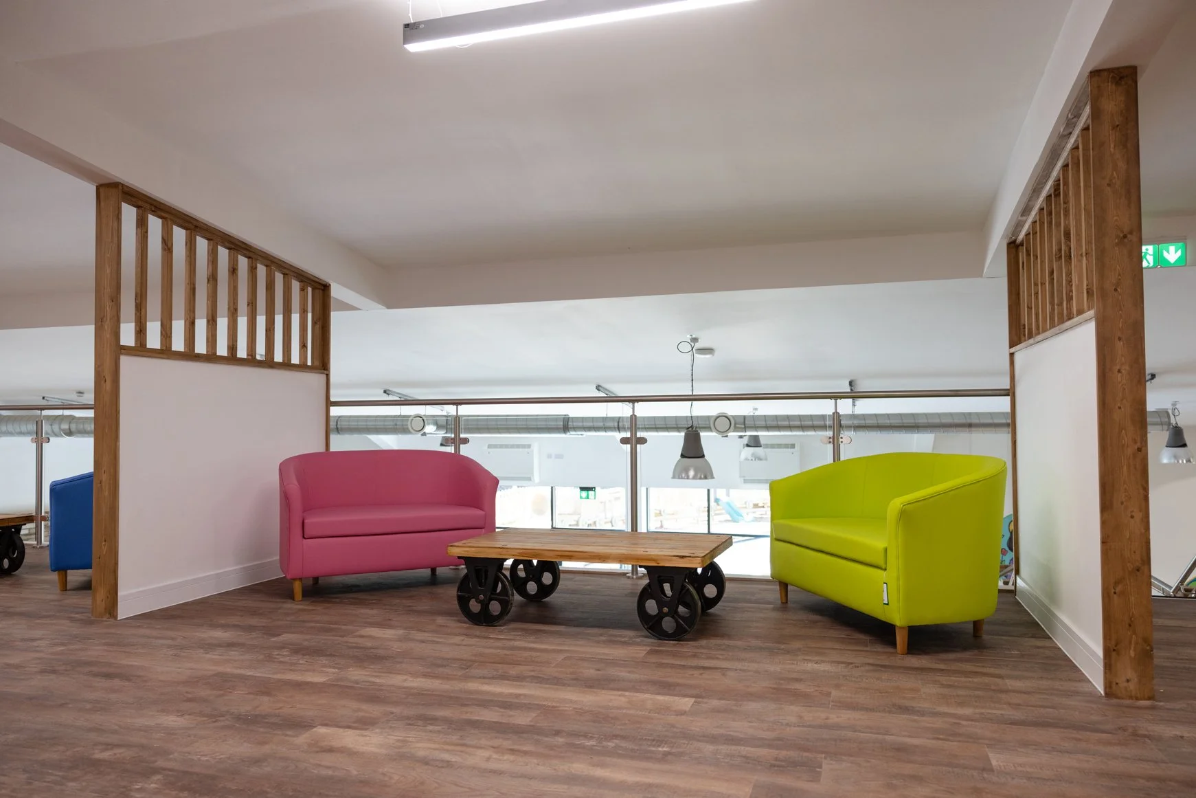 Interior view of a modern lounge area with colorful sofas, a low wooden table on wheels, and wooden accents, overlooking large windows.