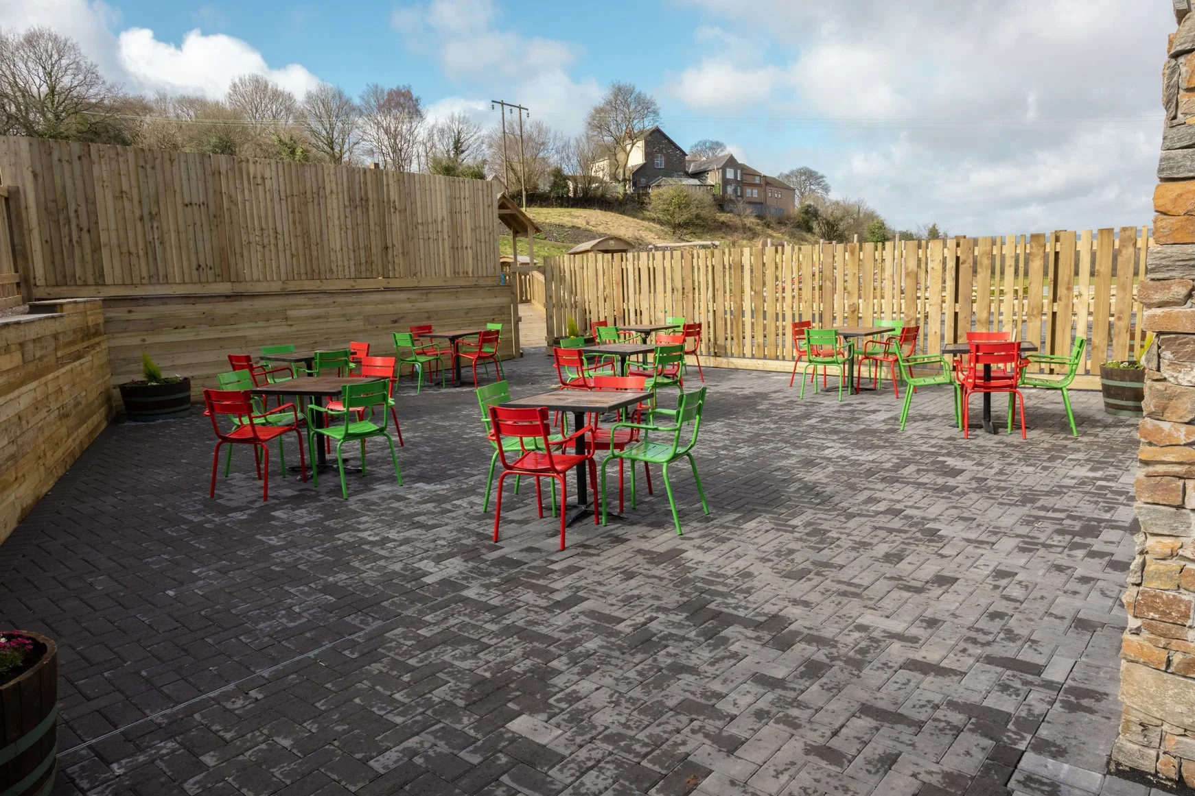 Outdoor patio with multiple red and green metal chairs and tables on dark brick pavers, surrounded by wooden fencing and a few potted plants, with houses and trees on a hill in the background.