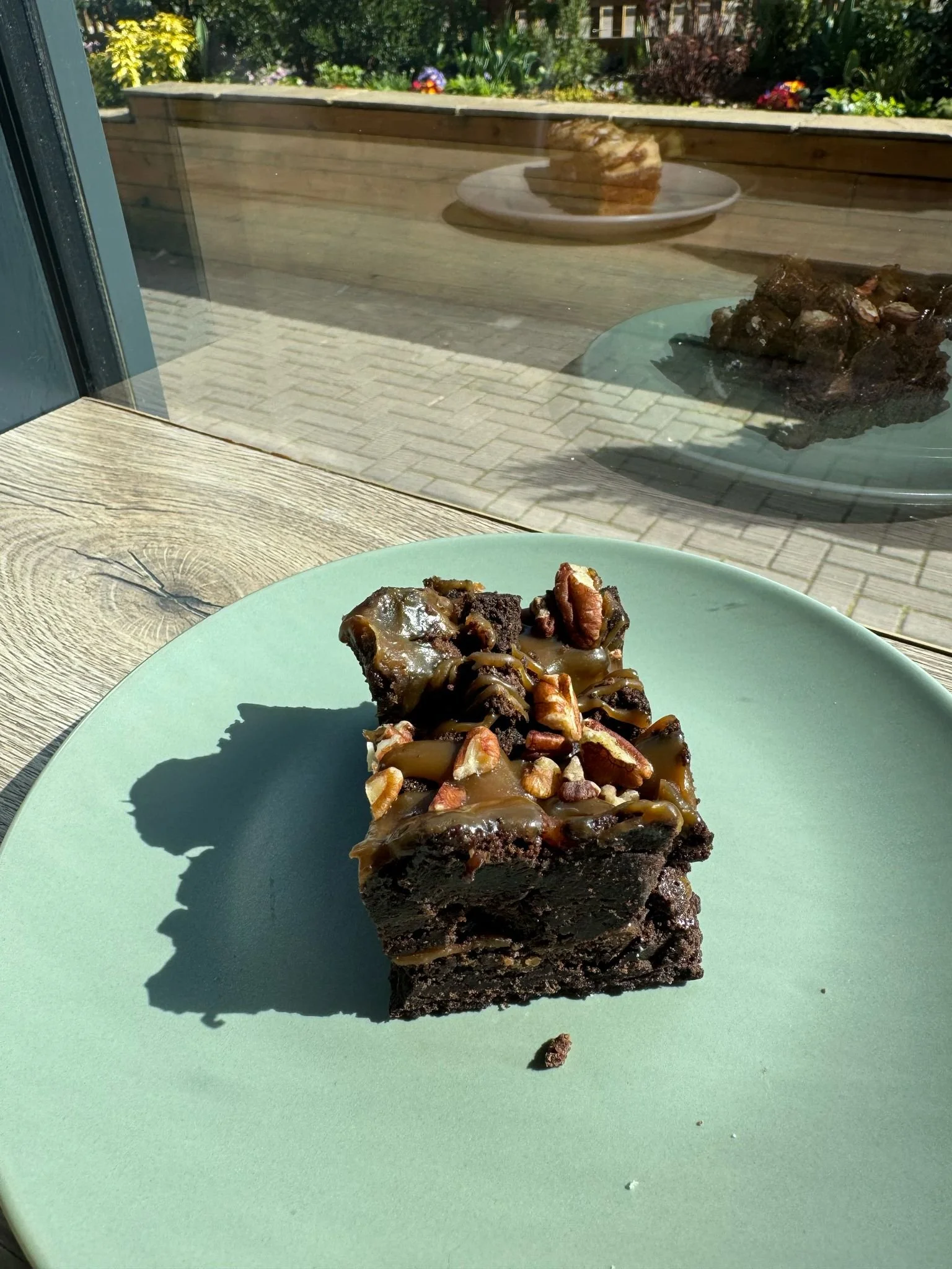 A close-up of a chocolate brownie topped with caramel and chopped nuts on a light green plate, sunlight casting a shadow, outside view of a patio with another plate of brownie and pouring rain reflected on the window.