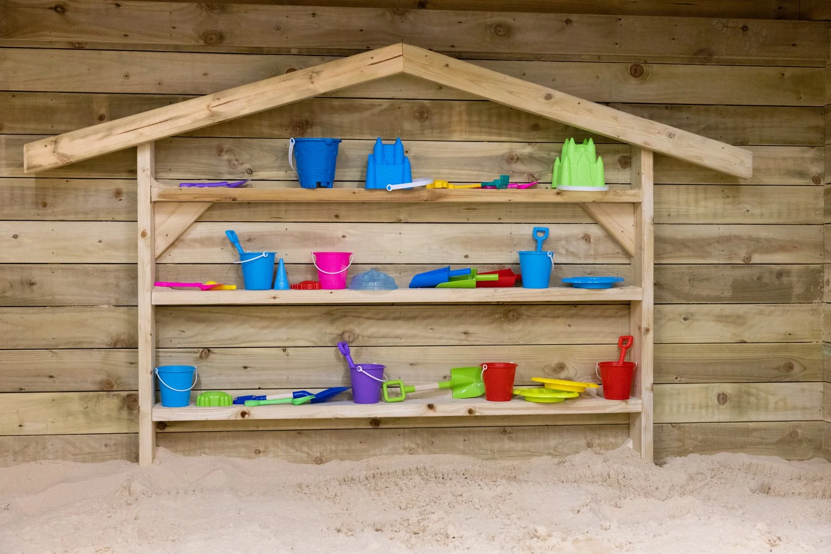 Colorful plastic beach toys on wooden shelves against a wooden wall, with sand in the foreground.