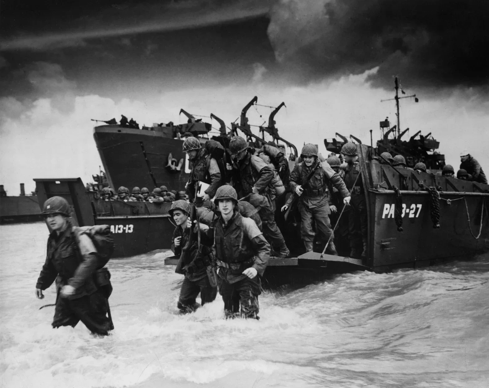 Black and white photograph showing soldiers disembarking from an amphibious assault vehicle into the water, with a military ship in the background under dark, cloudy skies.
