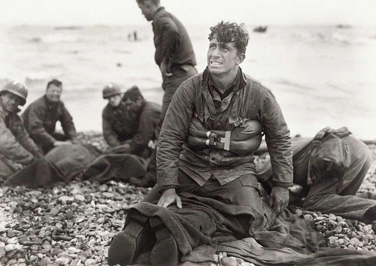 A black and white photograph of a group of soldiers on a rocky shoreline. One soldier is kneeling in the foreground, looking distressed, with others in the background attending to a wounded or fallen soldier.