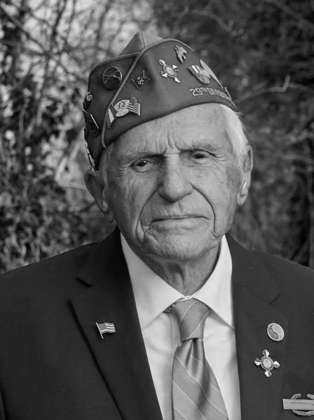 Elderly man in a suit and military hat holding a framed military photo and wearing decorated medals standing outdoors.