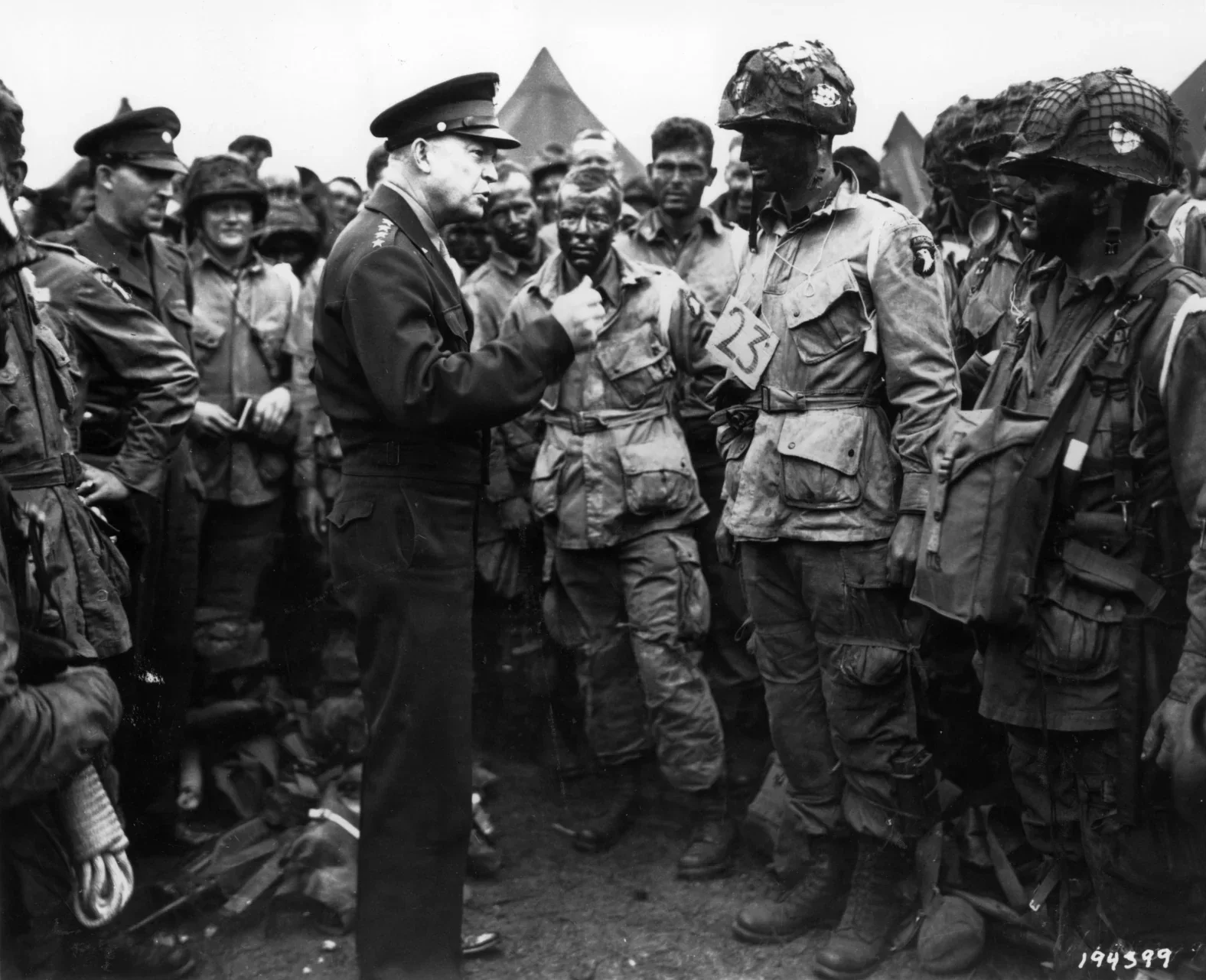 Black and white photo of a military officer speaking to soldiers in uniform, with other soldiers and tents in the background.