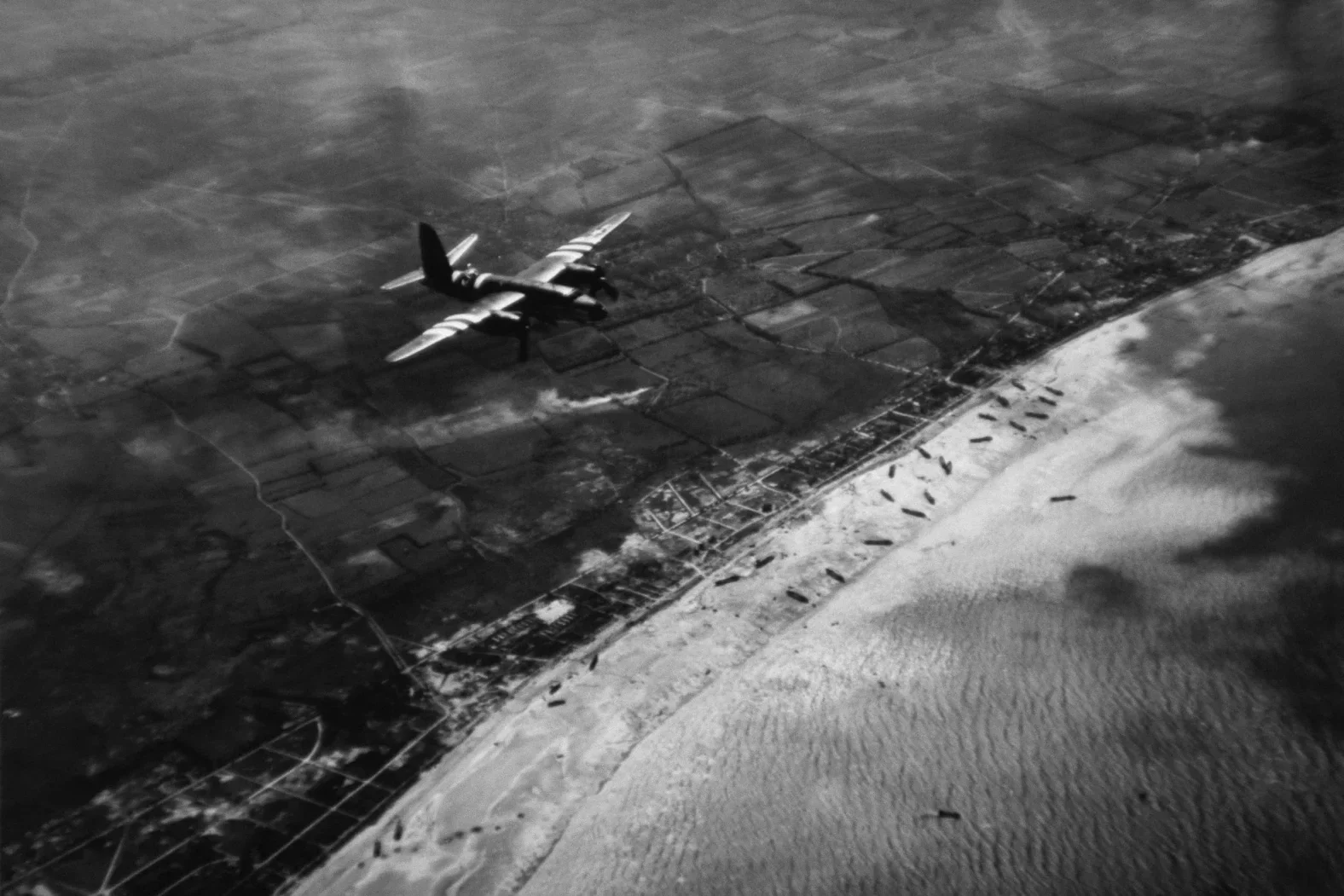 Black and white aerial photograph of a military airplane flying over a coastal landscape with fields, roads, and ships along the shoreline.