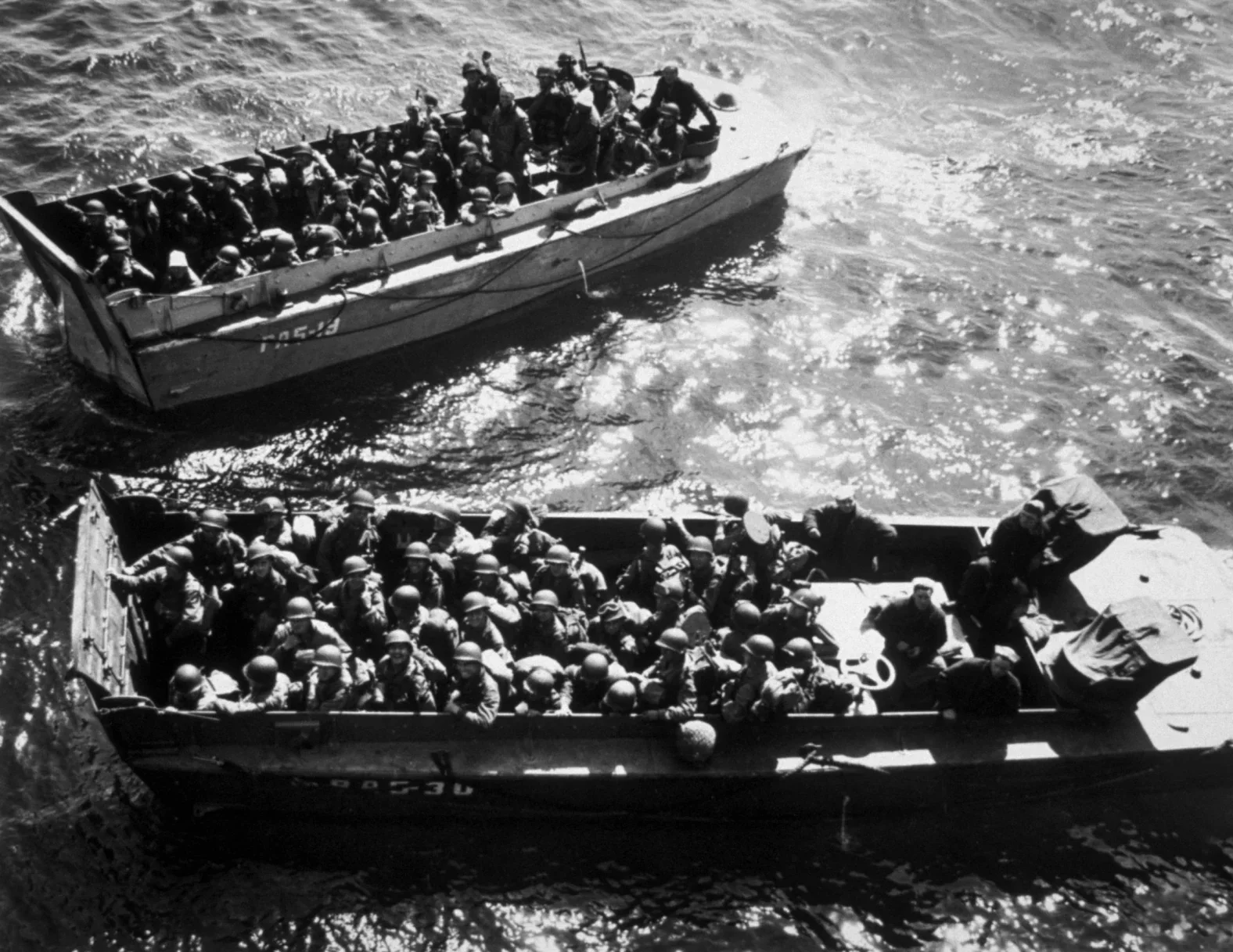 Black and white photograph of two military boats filled with soldiers sailing on the water.