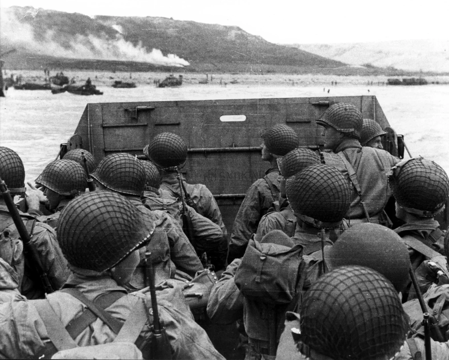 World War II soldiers in helmets, seated in a landing craft preparing for amphibious assault, with battleships and smoke in the background.