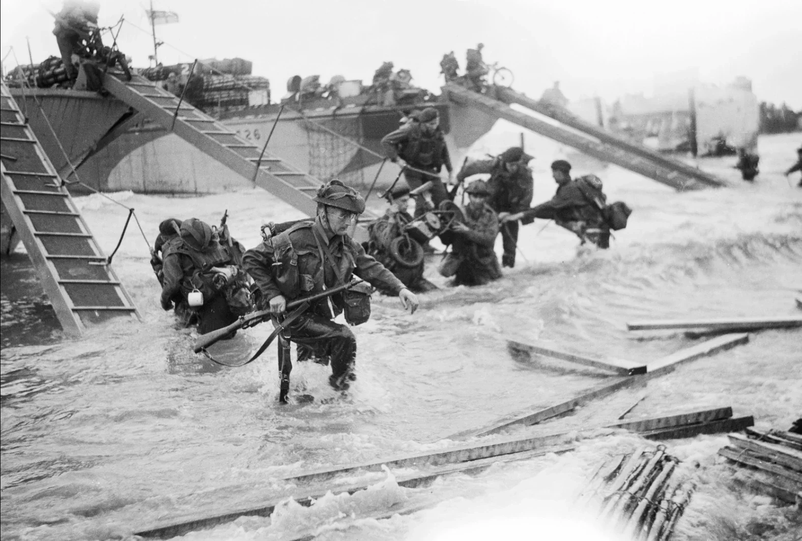 World War II soldiers wading through floodwaters, crossing a washed-out bridge with damaged wooden planks, during a military operation.