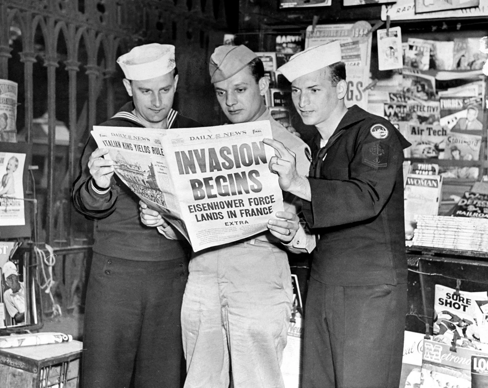 Three men in U.S. Navy uniforms standing together in a store, reading a newspaper with headlines about a military invasion in France, from the World War II era.