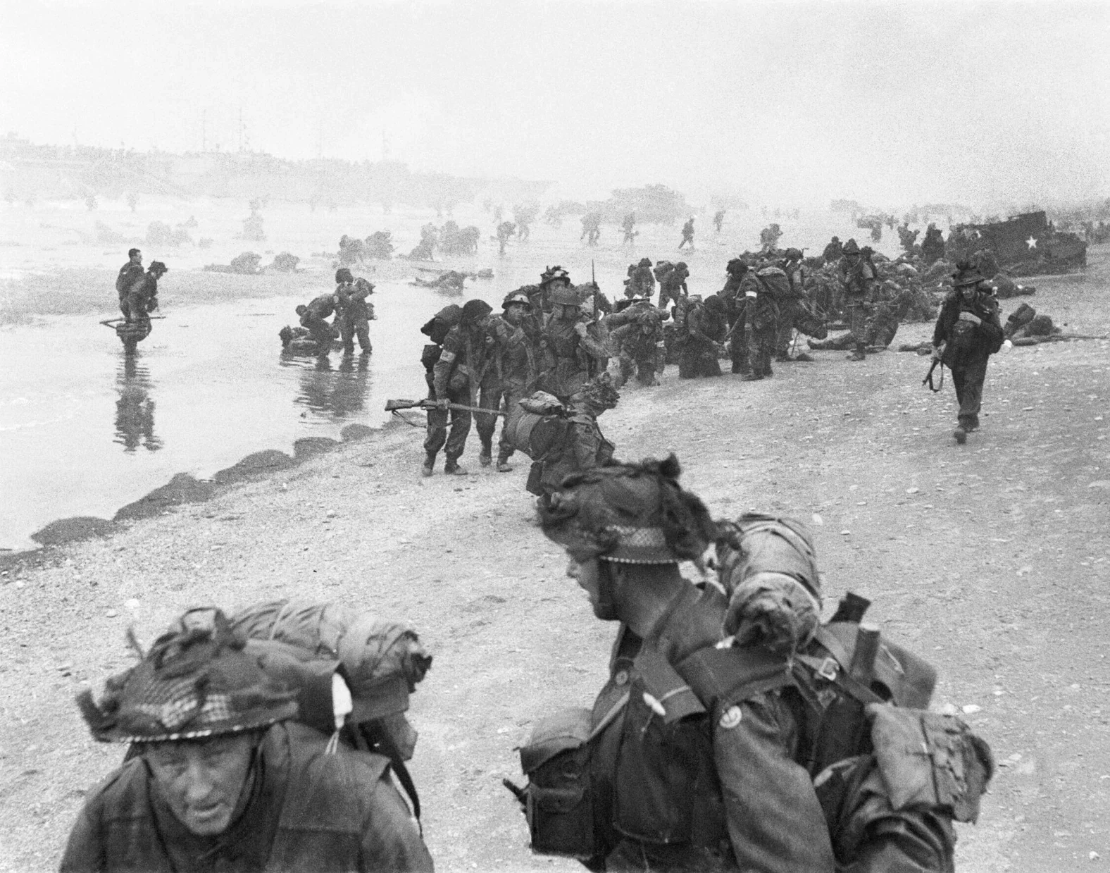 Black and white historic photograph of soldiers crossing a river during wartime, with some wading through water and others on the bank carrying gear.