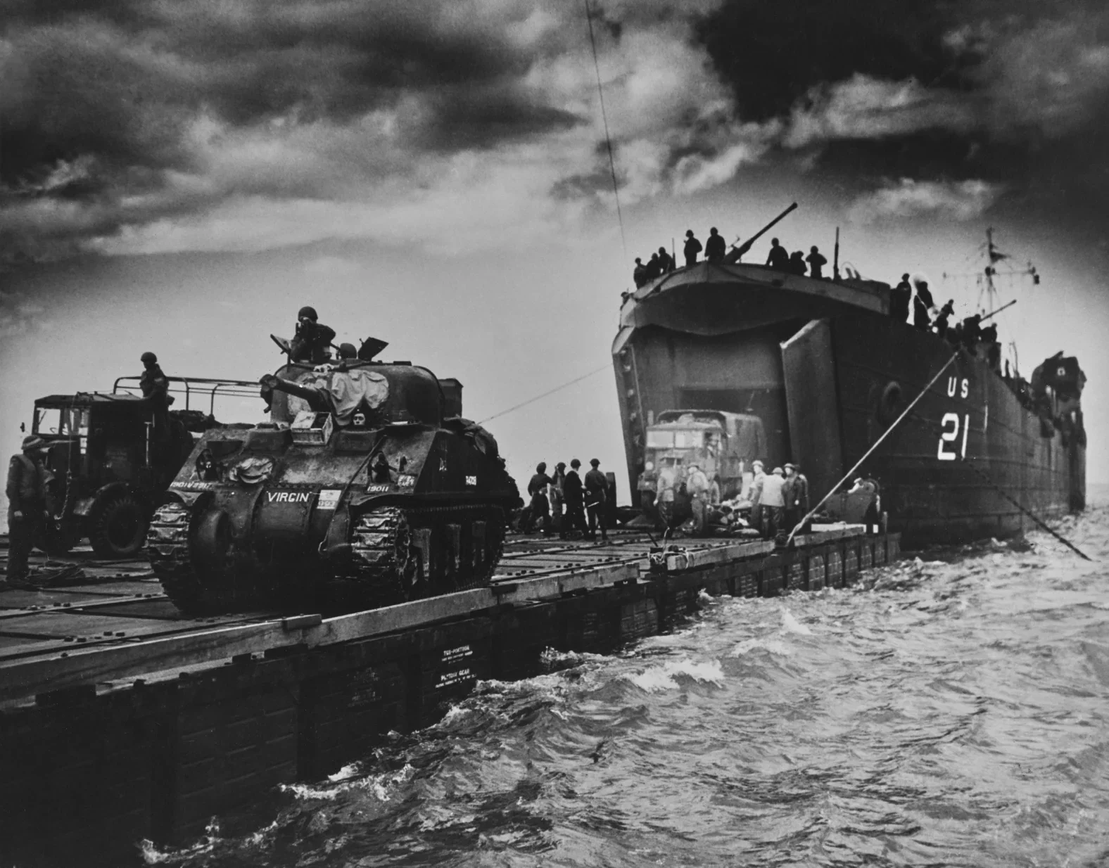 Black and white photo of soldiers and military vehicles on a ship, with a tank on the deck and soldiers disembarking to a boat, maritime setting with cloudy sky.