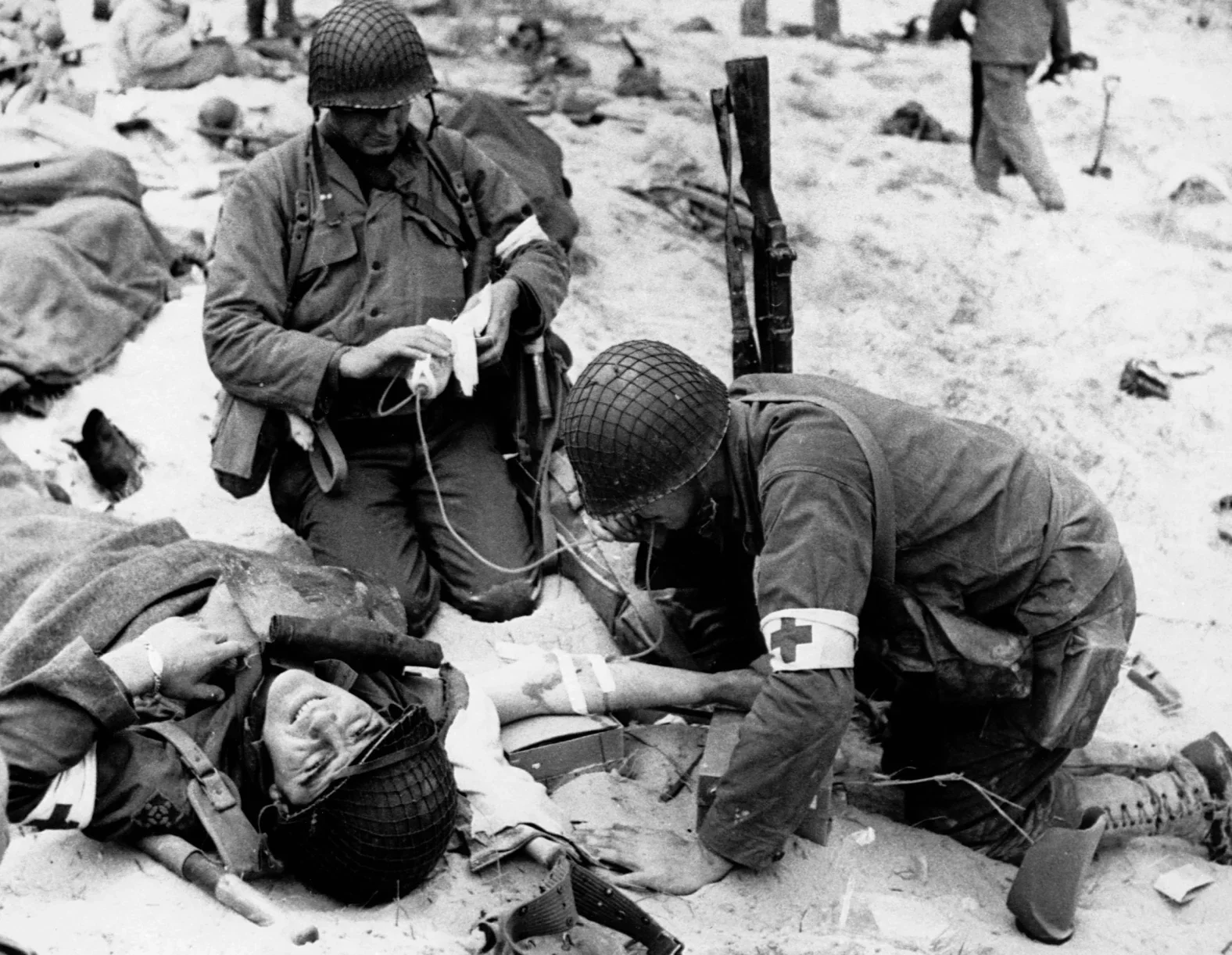 Two soldiers in World War II uniforms tending to a wounded soldier lying on the ground, with debris and rubble scattered around in a battlefield.