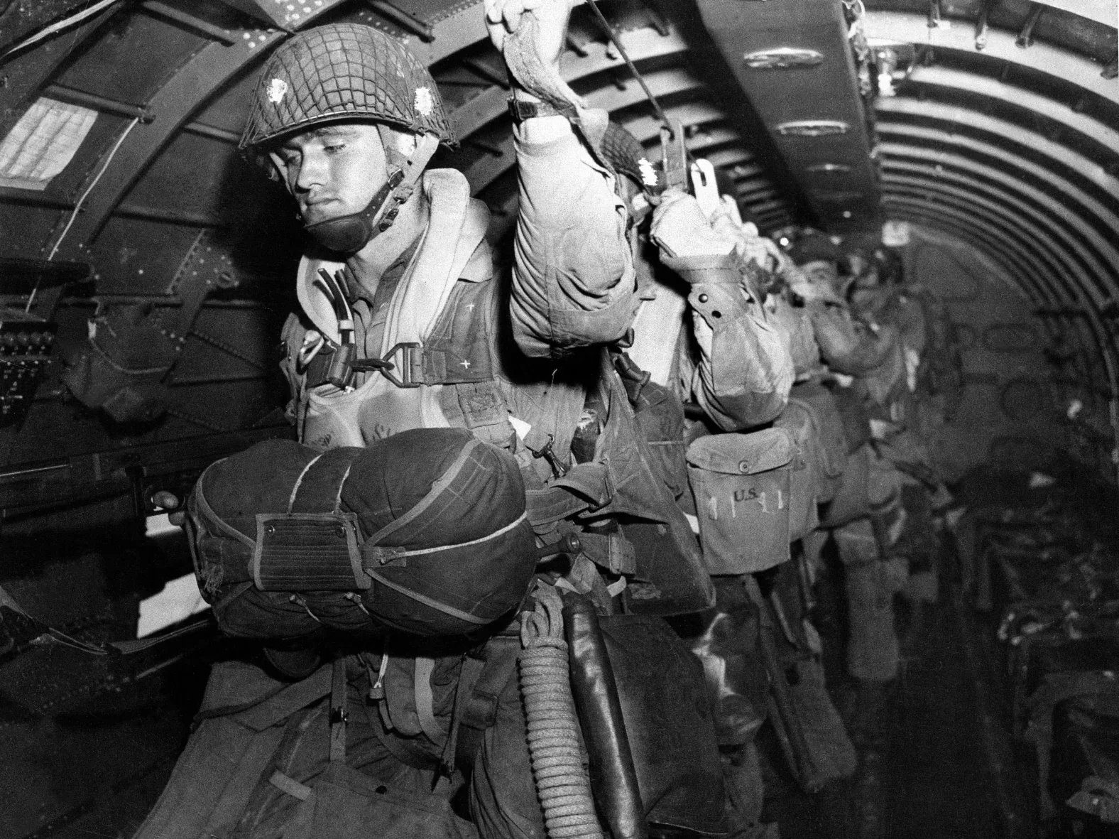 A black and white photograph of soldiers inside a military aircraft, with one soldier in the foreground wearing a helmet, gear, and holding a wire or tool, while others are arranged in rows behind him.