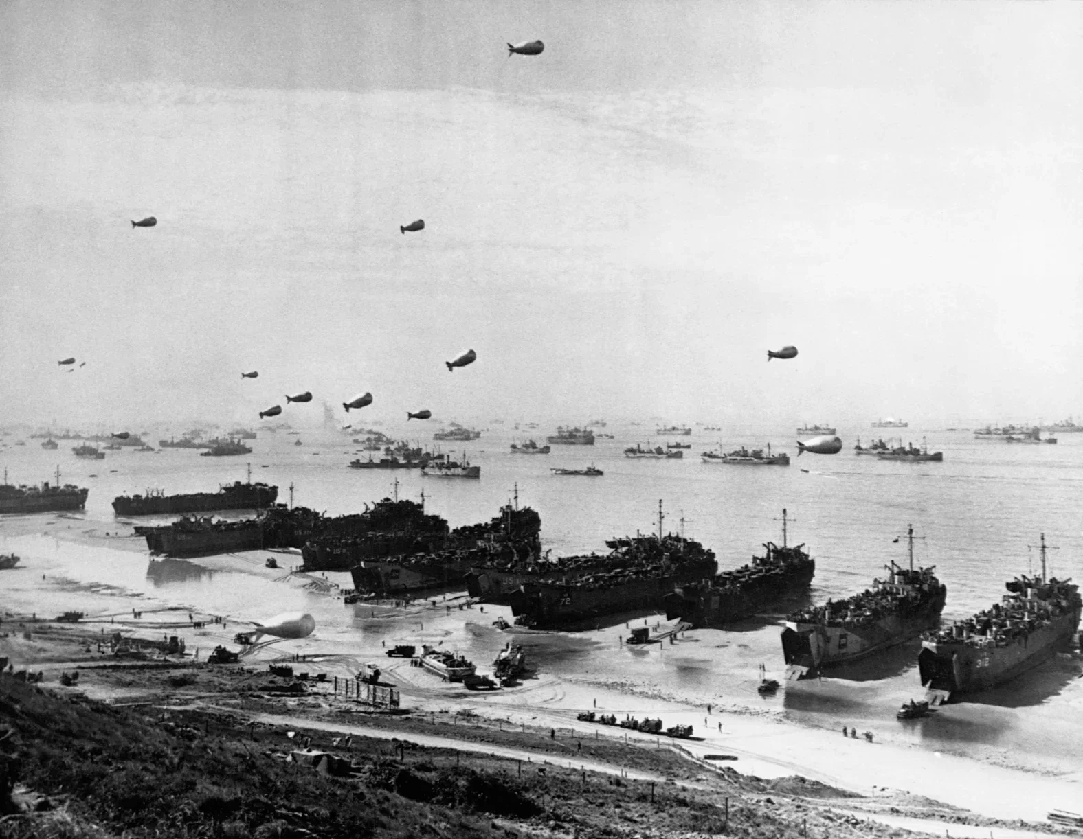 Historical black and white photograph of a naval fleet, showing numerous ships in the water and several large airships flying overhead with a coastline in the foreground.