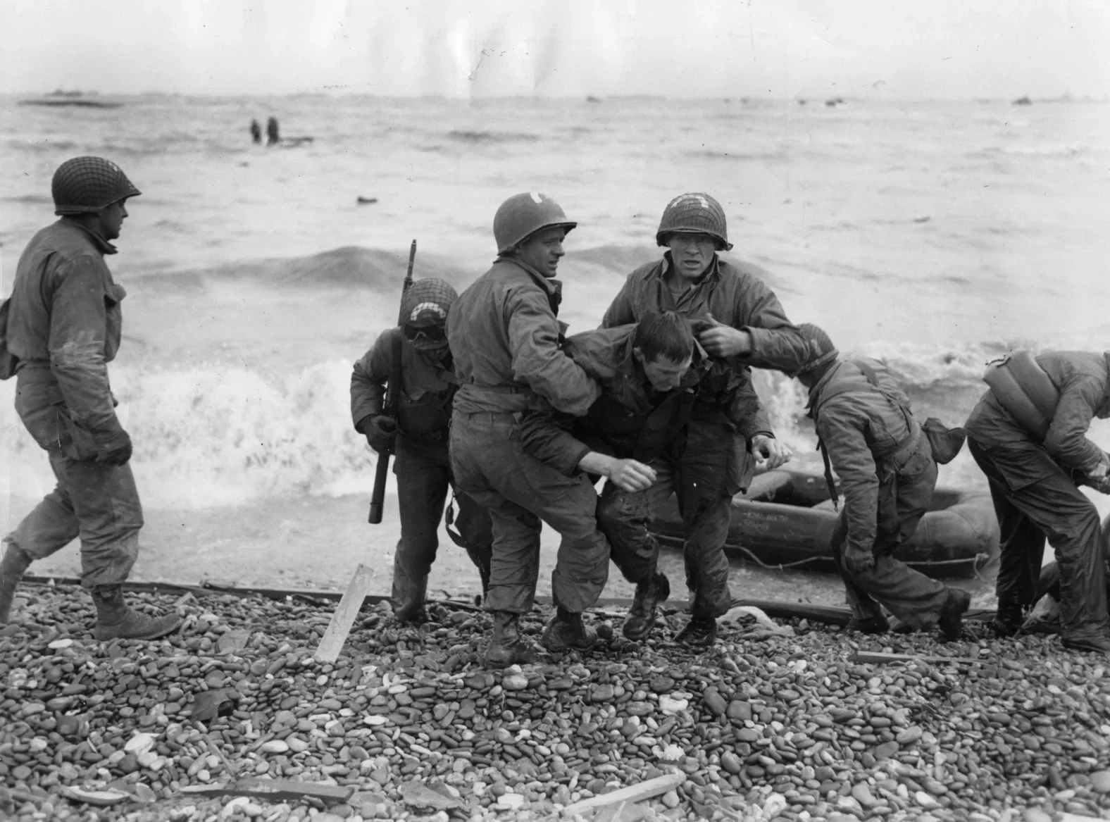 A group of soldiers in military gear assisting a wounded comrade onto a boat on a pebbled beach with the ocean in the background.