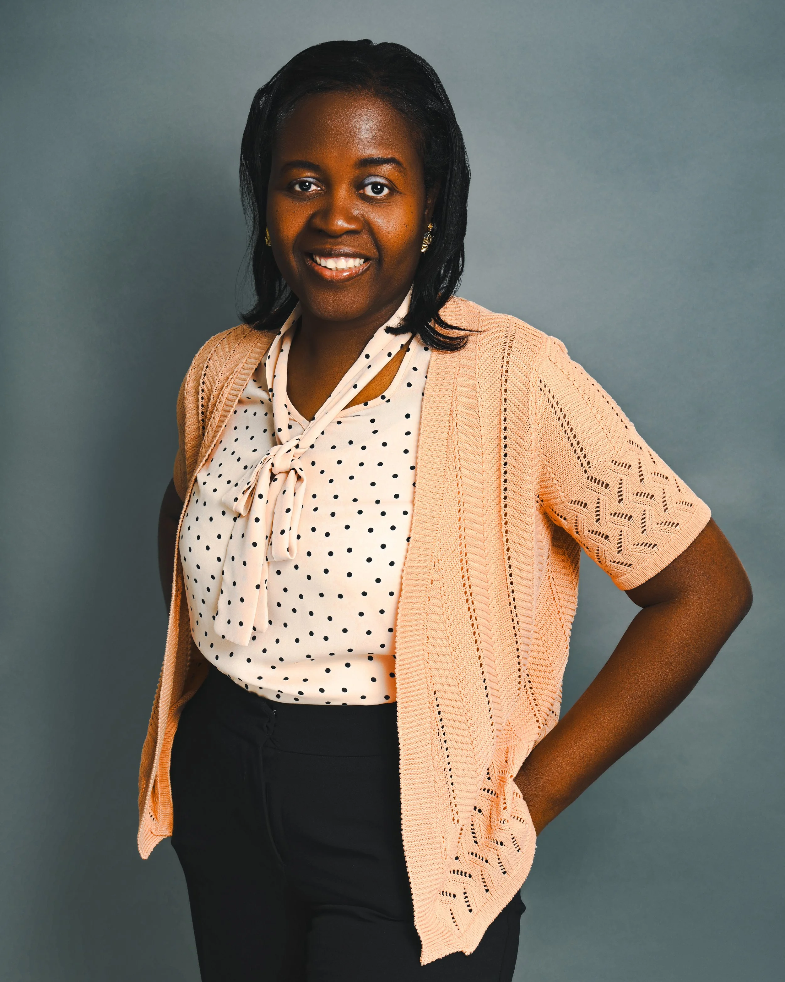 A smiling woman standing against a grey backdrop, wearing a peach knit cardigan over a polka-dot blouse and black pants, posing confidently with her hands in her pockets. She is the founder of Manna Help, a non-profit supporting families facing food 