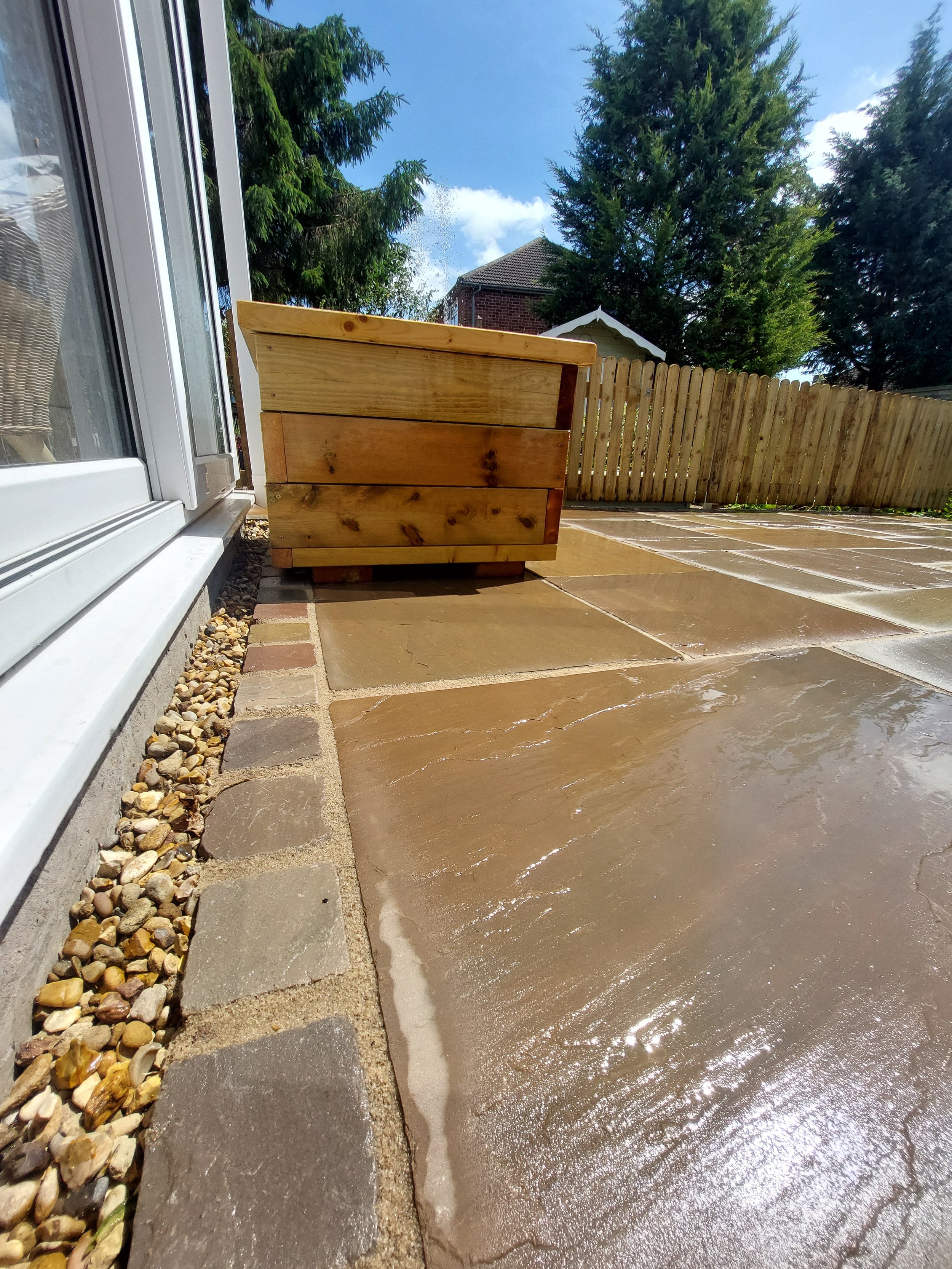 A wooden outdoor planter on a wet stone patio, with a sliding glass door on the left and a wooden fence in the background, under a partly cloudy sky.