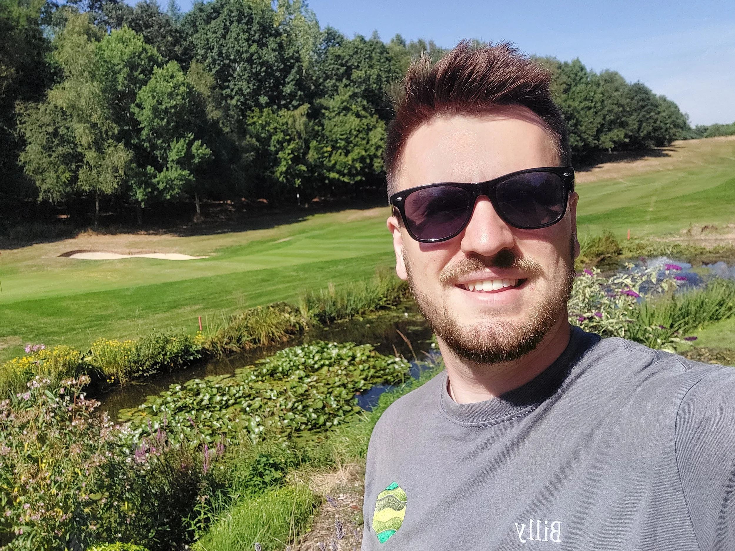 A man with sunglasses smiling outside near a pond with water lilies, a golf course, and trees in the background.