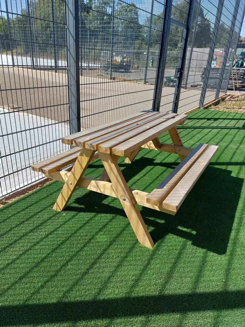 Wooden picnic table and bench on green artificial turf outside enclosed by metal fence.