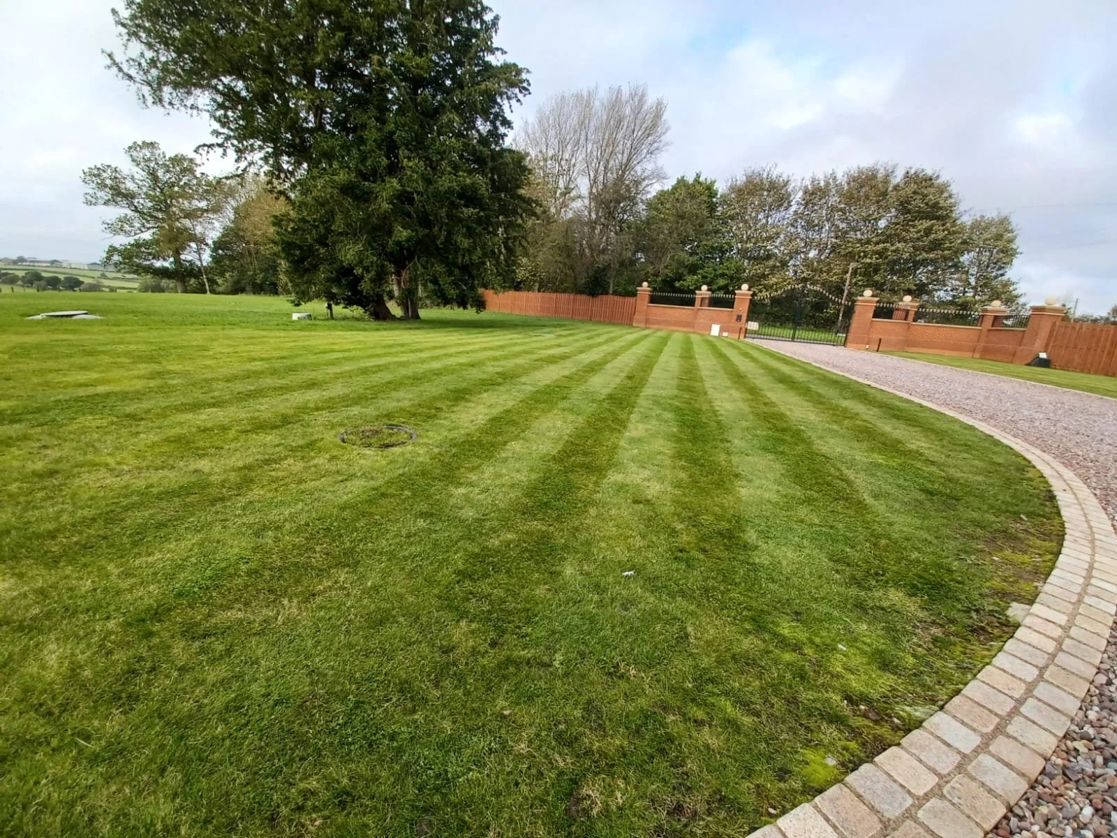 Well-maintained lawn with striped grass, trees, a gravel driveway, and a brick wall with iron fencing in the background.