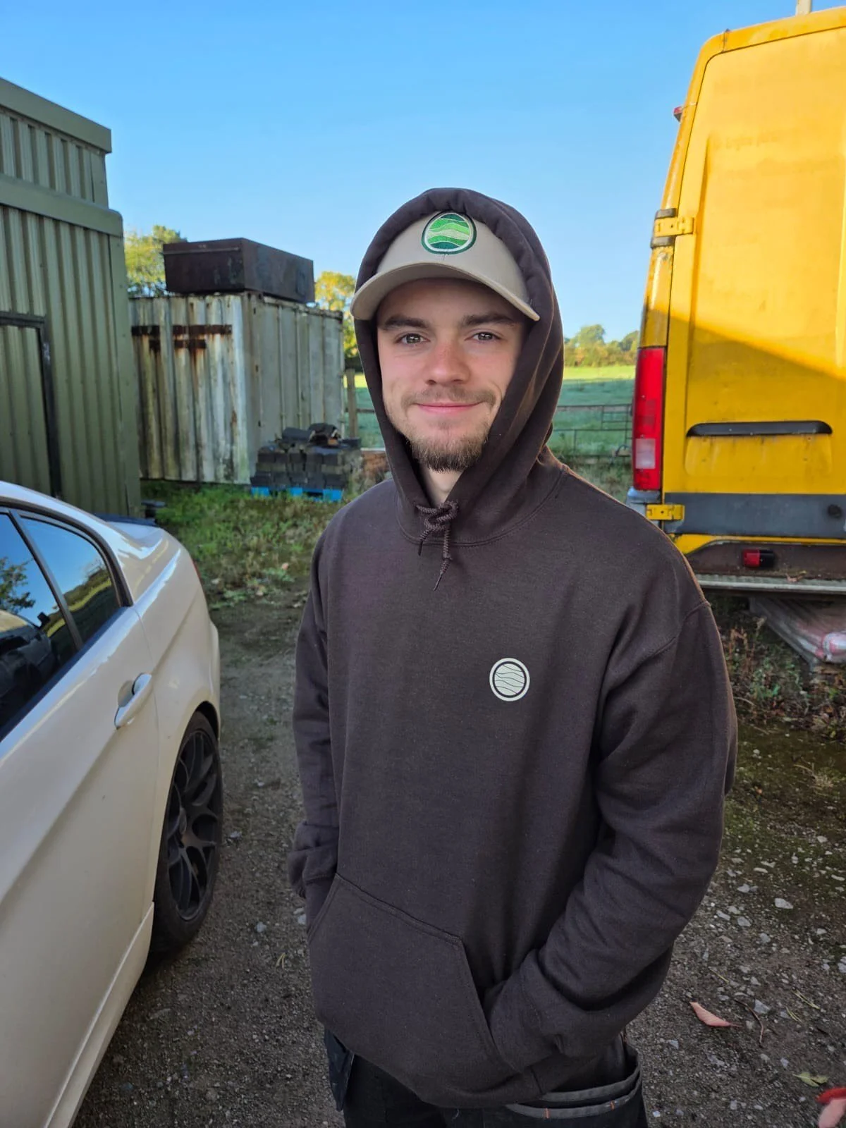 Young man standing outdoors in a hoodie and cap, smiling, with vehicles and storage containers in the background.