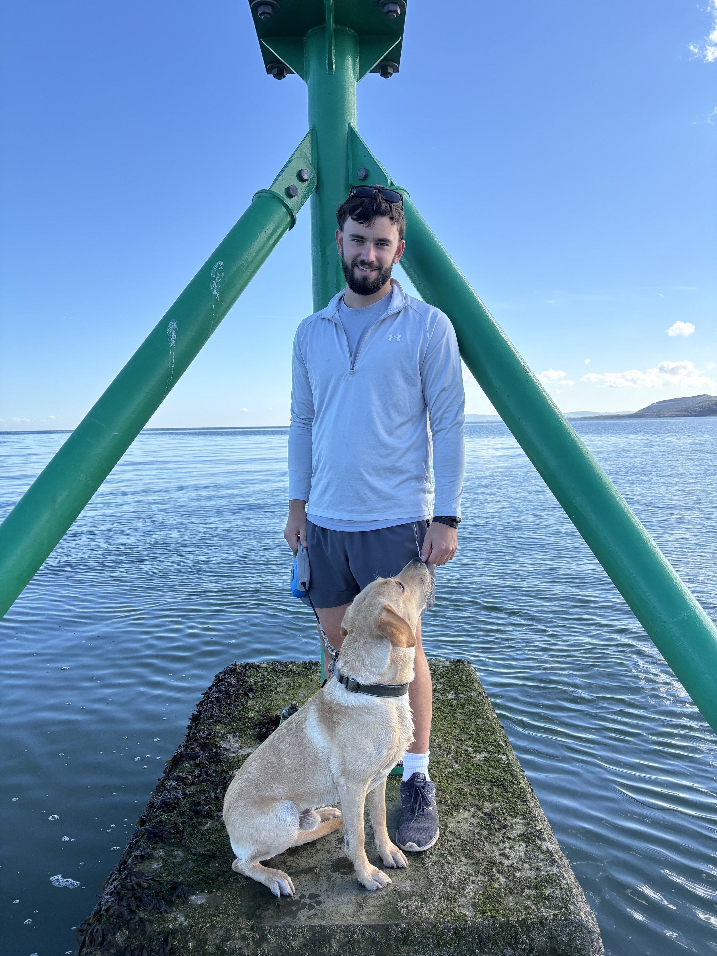 A man with a beard and sunglasses on his head standing on a moss-covered concrete pier by the water, holding a blue retractable leash attached to a tan dog with floppy ears. The background features a clear blue sky with a few scattered clouds and distant hills.