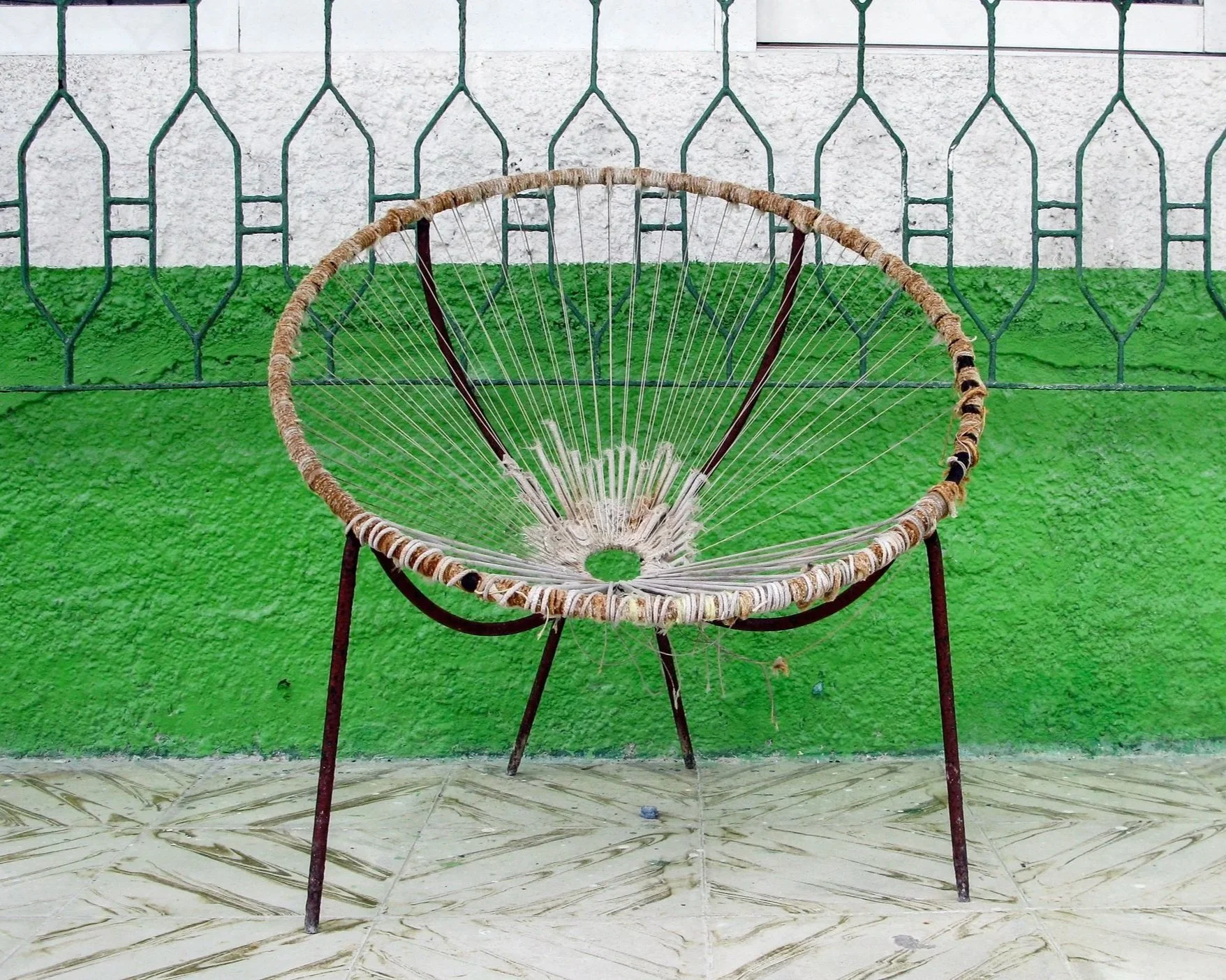 A chair made from a circular woven frame with a spider web in the center, leaning against a green painted wall with a white upper section and a decorative metal fence above.