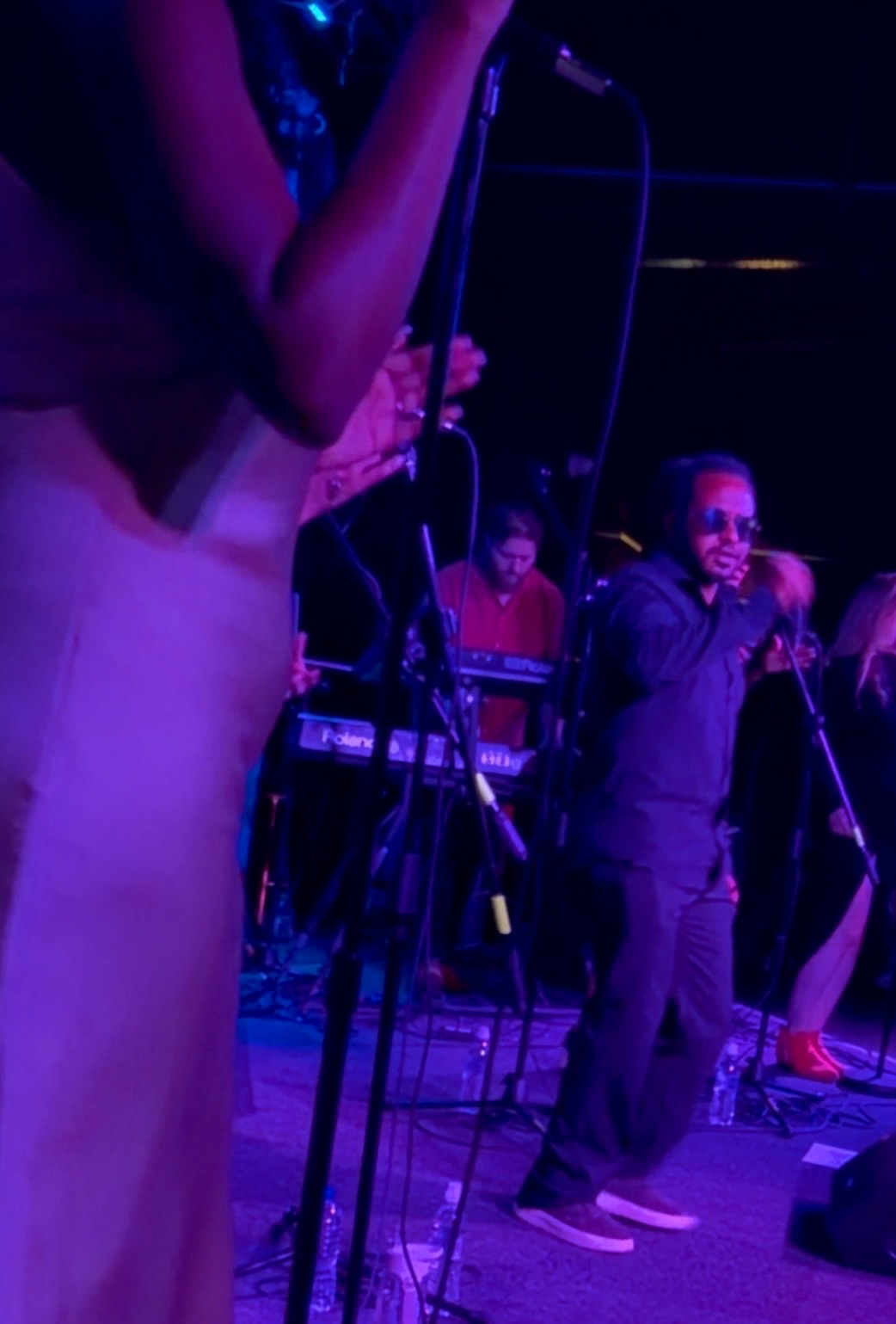 Musicians performing on stage in a dark setting with purple lighting, with a male singer in sunglasses and a dark jacket standing in front of microphones and keyboard players visible in the background.