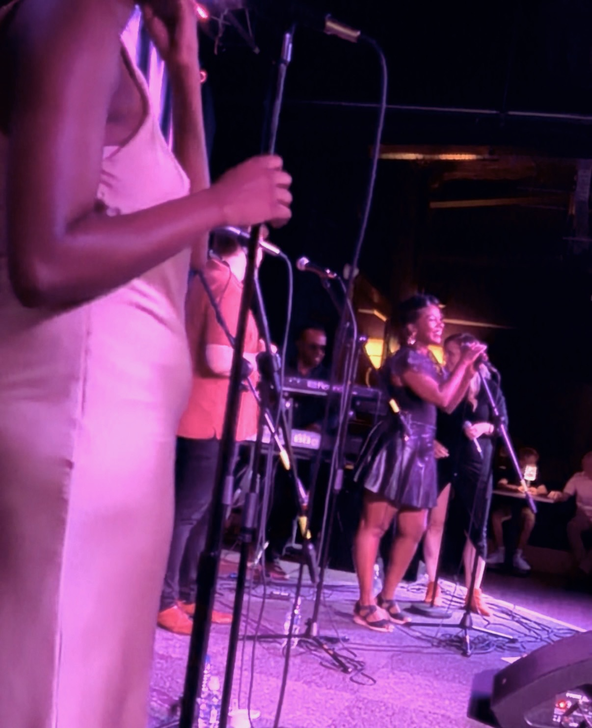 A group of vocalists singing on stage with microphones, surrounded by music equipment, with an audience sitting at tables and on the dancefloor in the background.