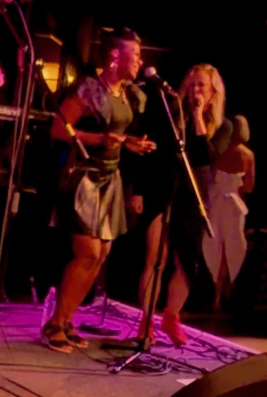 Two women standing on a stage singing, colorful stage lighting, and musical equipment in the background, possibly performing or speaking at an event.