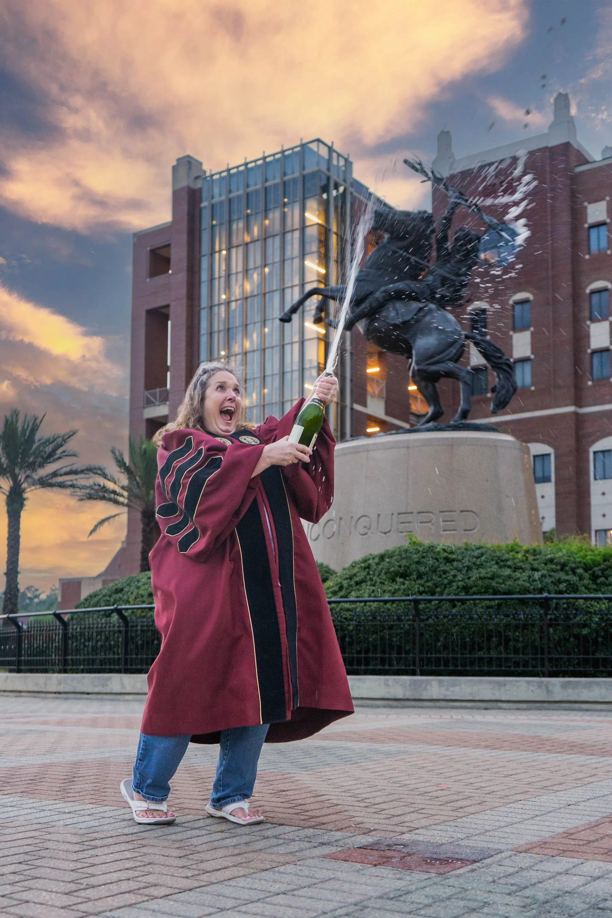 Female graduate student sprays champagne for her cap and gown photos with the Unconquered FSU statue in the background