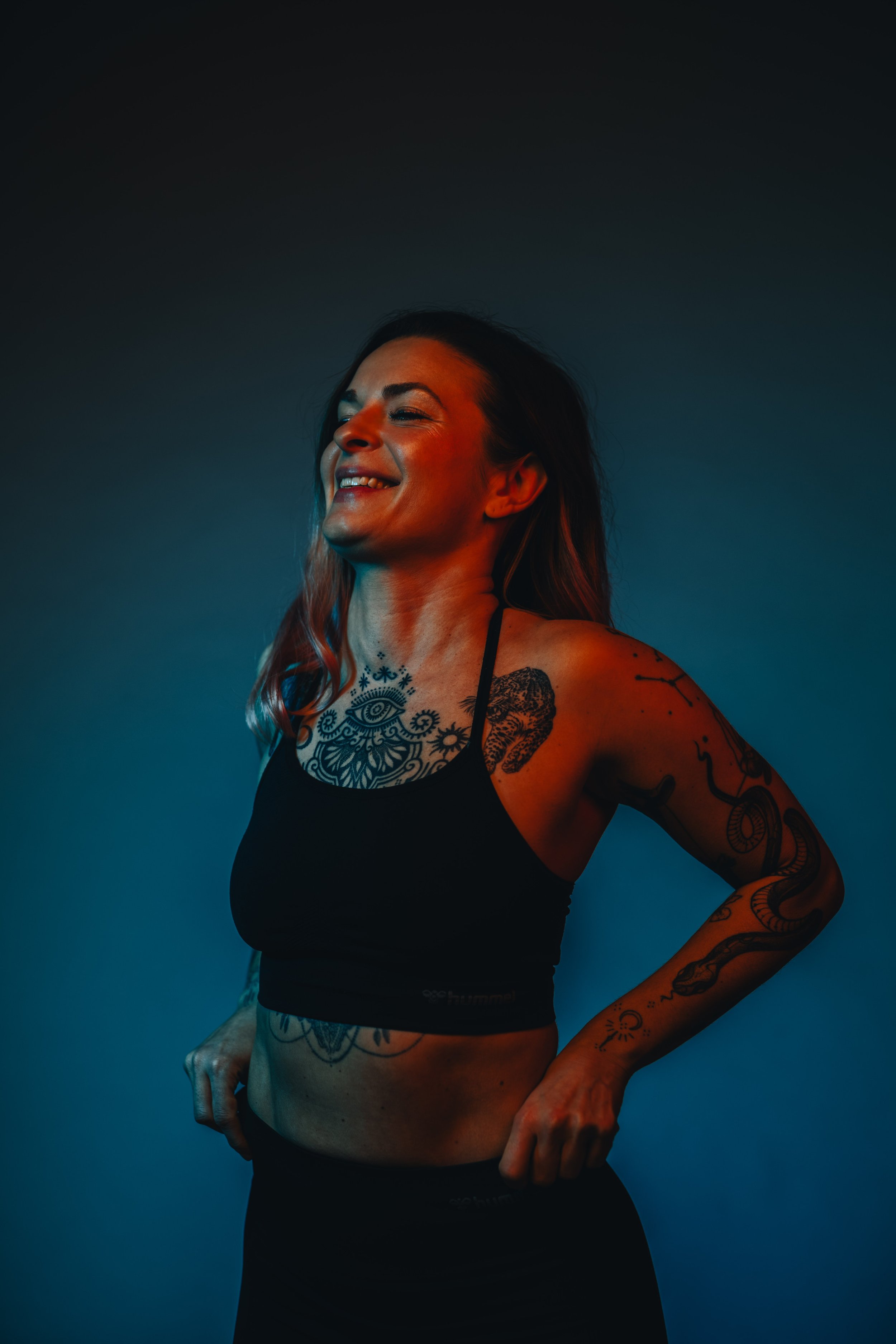 Eine lachende Frau mit Tattoos trägt eine schwarze Sport-BH, vor einem dunklen Hintergrund.