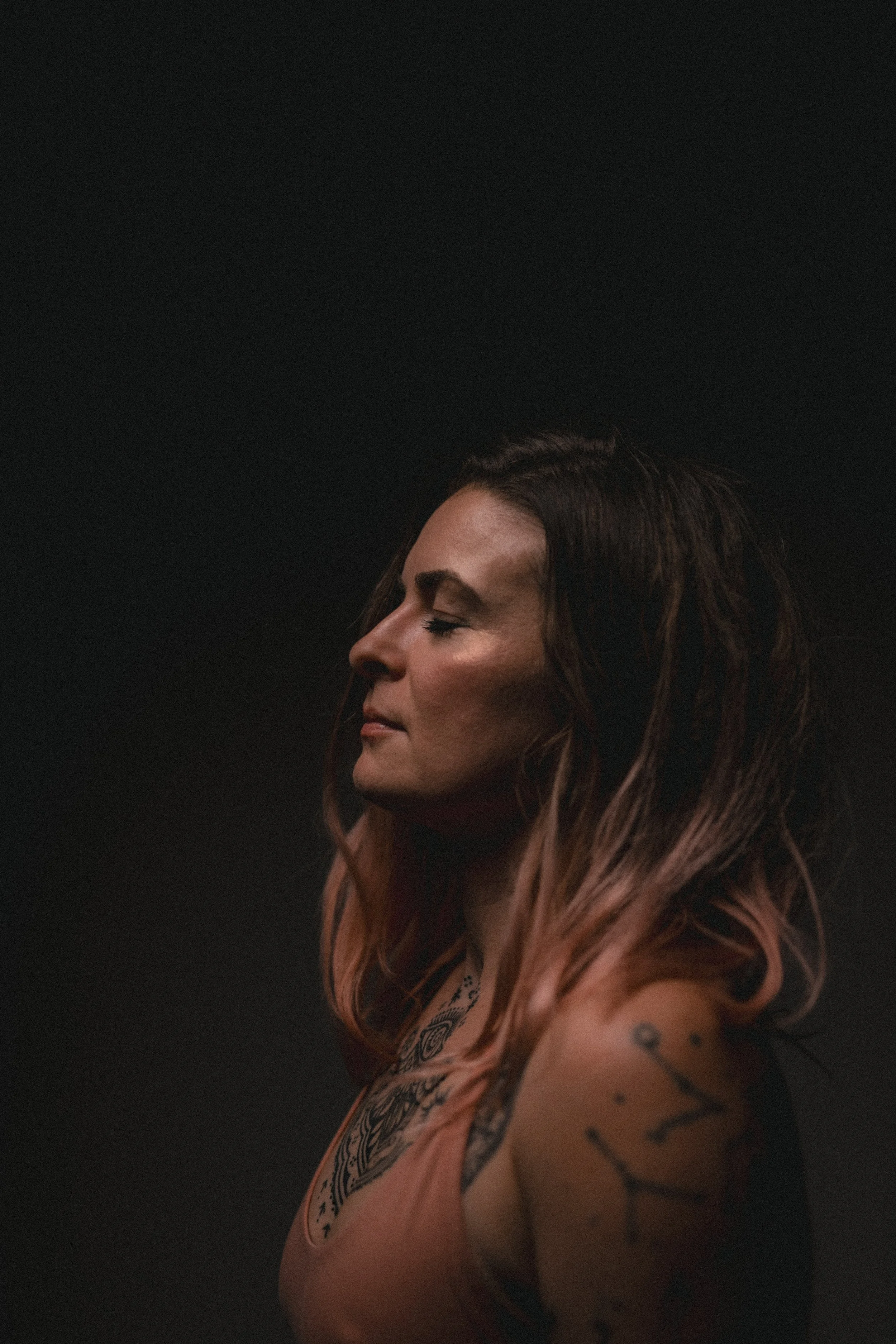 Porträt einer Frau mit langen braunen Haaren, Tattoos auf Arm und Schulter, seitlichen Blick, dunklem Hintergrund.
