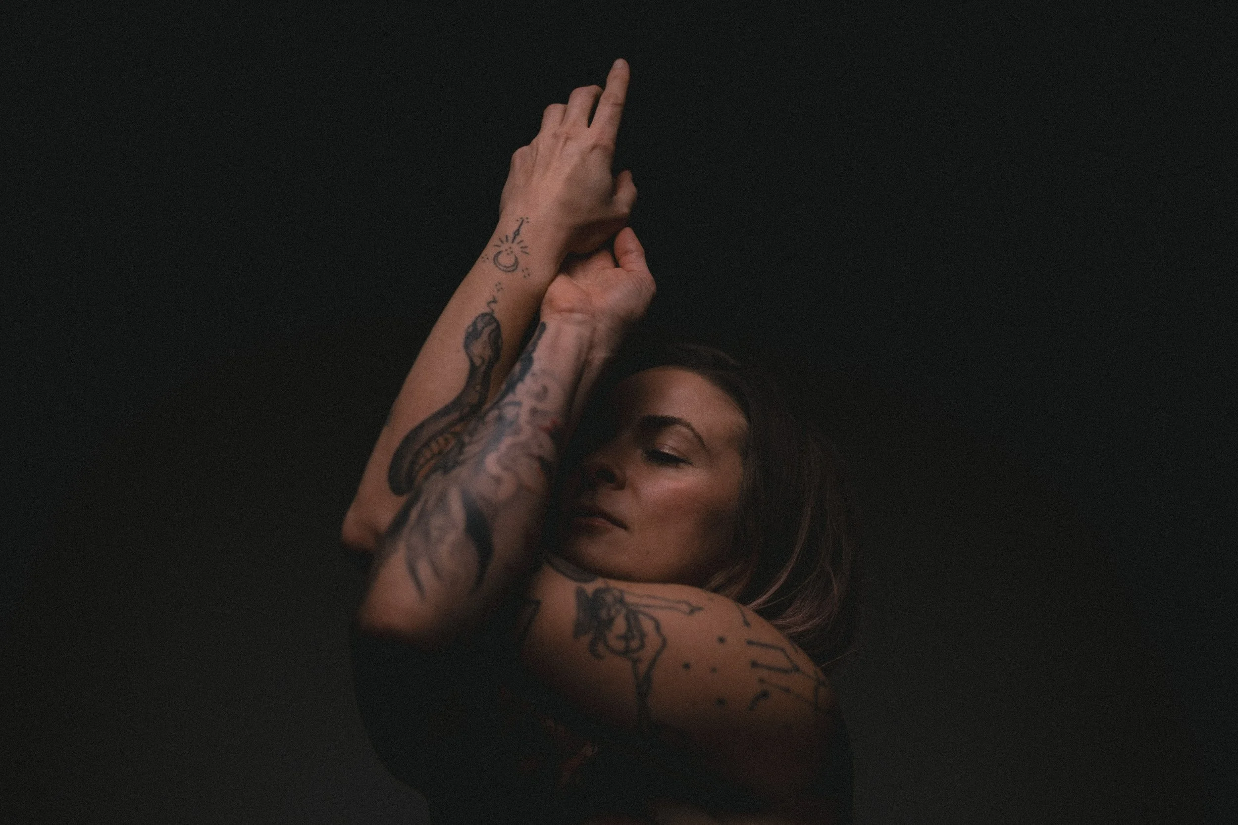 Junge Frau mit Tattoos, die sich mit den Händen über dem Kopf berührt, vor schwarzem Hintergrund.