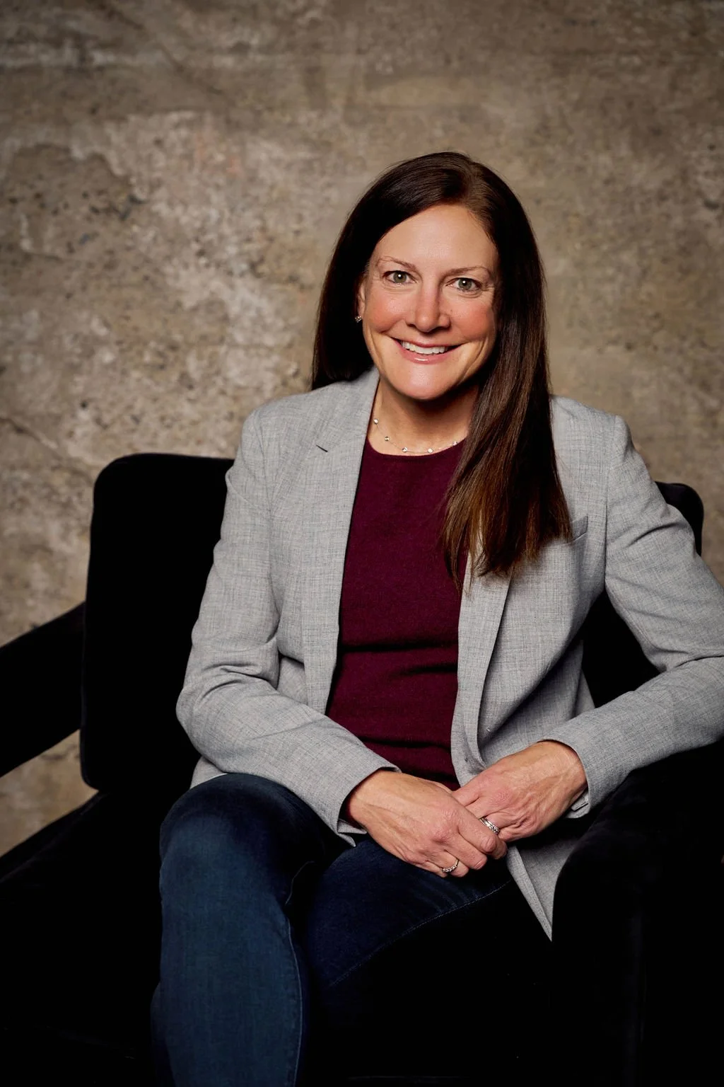 Woman seated in a gray blazer and purple top, smiling, with a textured background.
