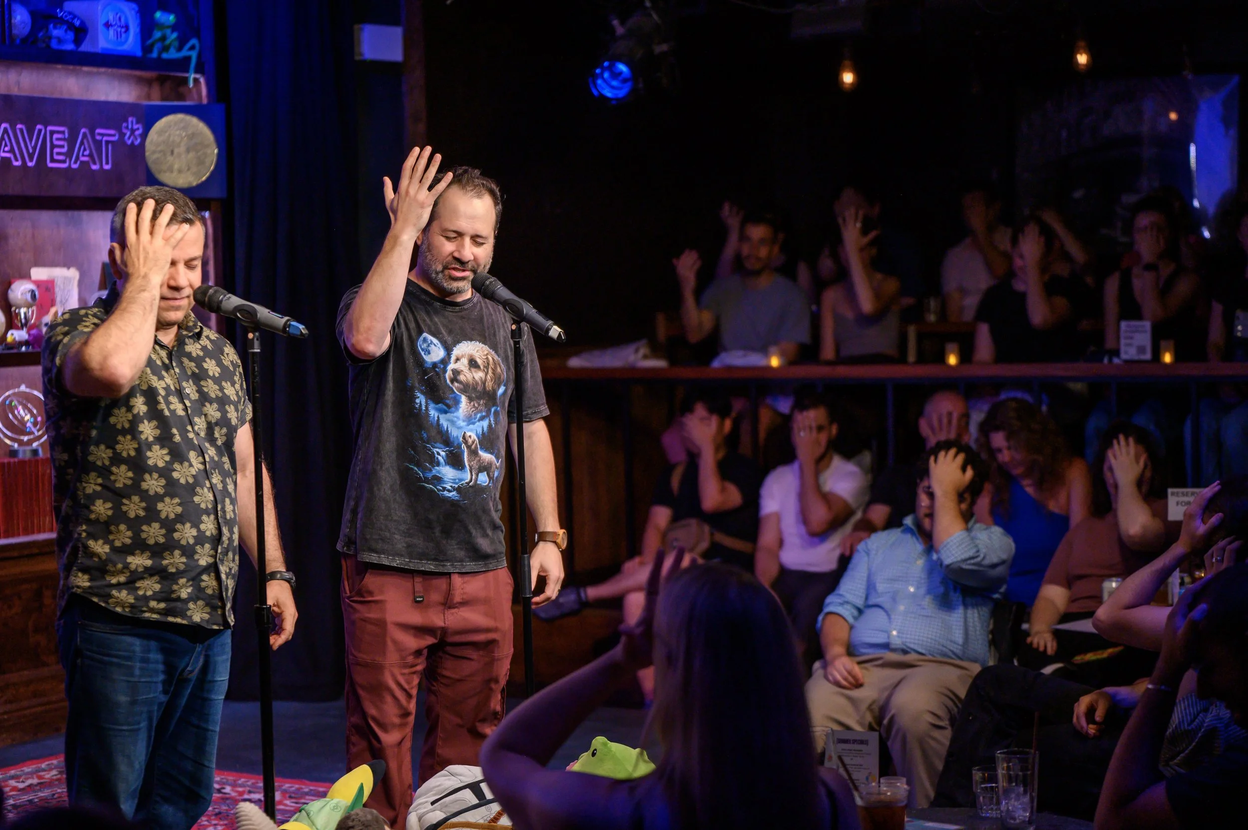 Co-hosts Jonah and Brady demonstrate face palm technique while audience mirrors in NYC at Caveat July 28th, 2026
