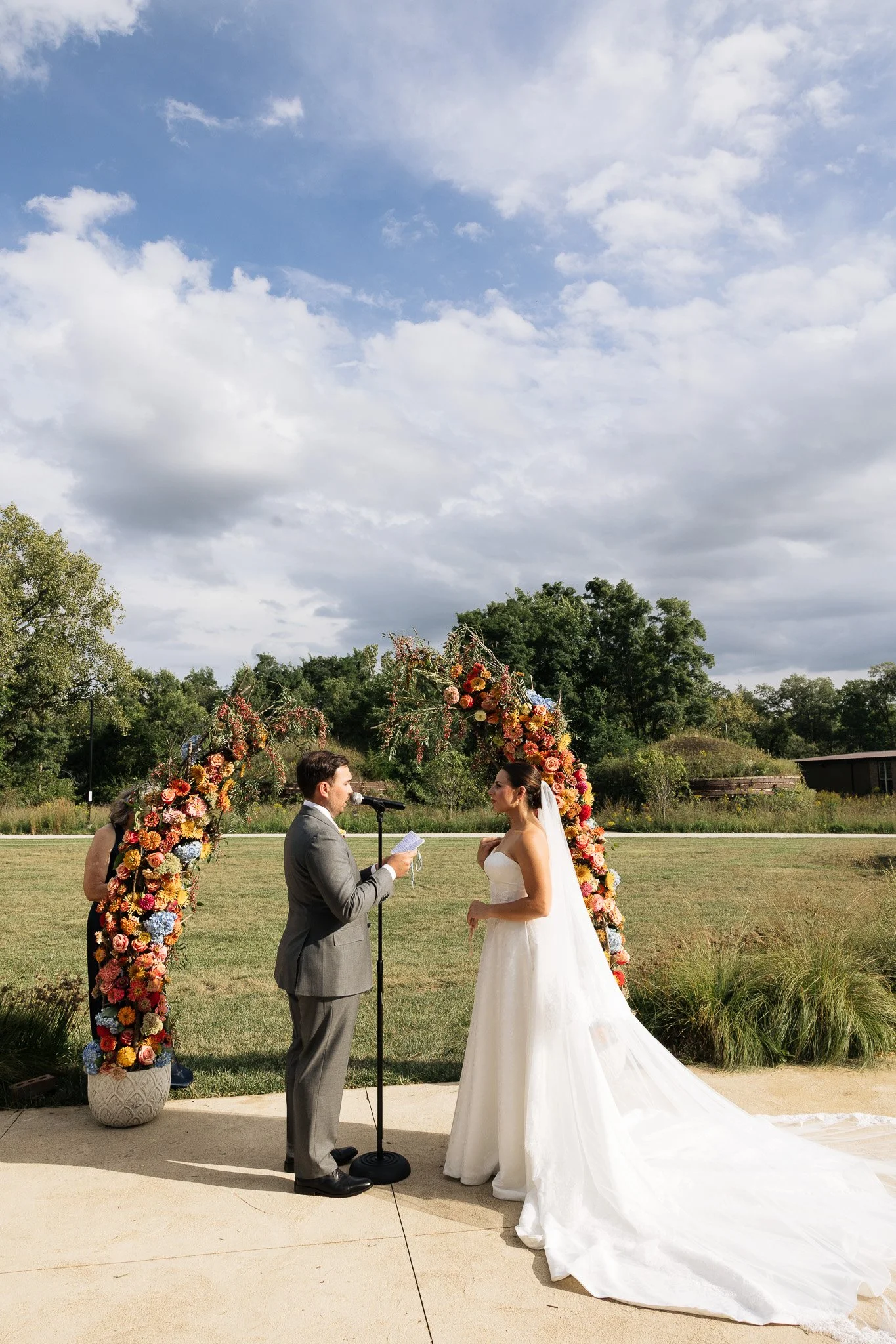 A bride in white standing across from her groom saying their wedding vows in front of a colorful floral arch