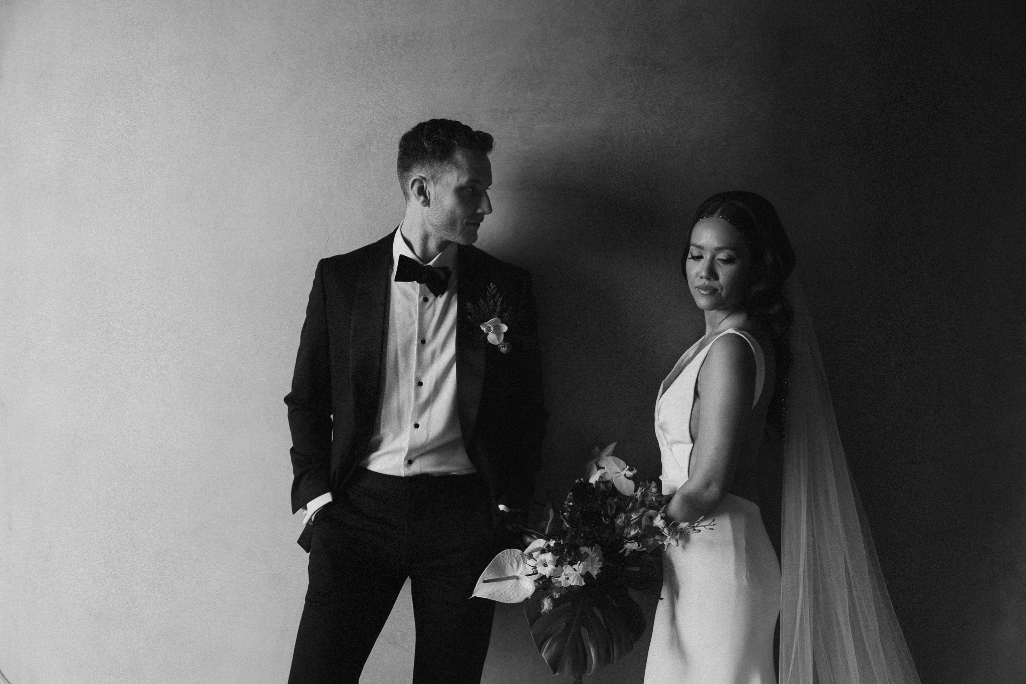 Editorial portrait in black and white of groom looking towards his bride against a wall.