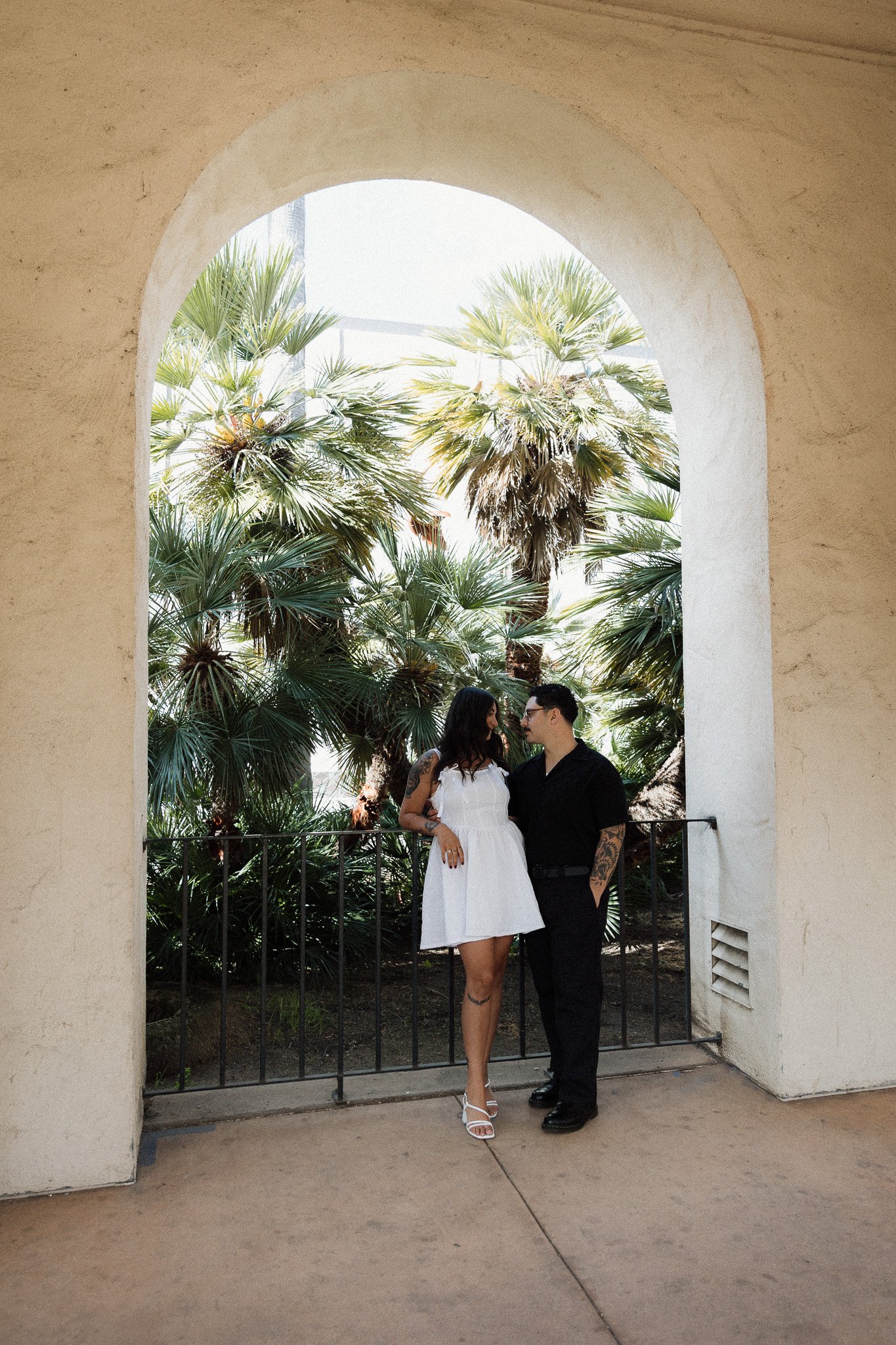 A couple standing underneath an arched structure with a background of palm trees, one woman wearing a white dress and heels, the man in a black shirt and pants.