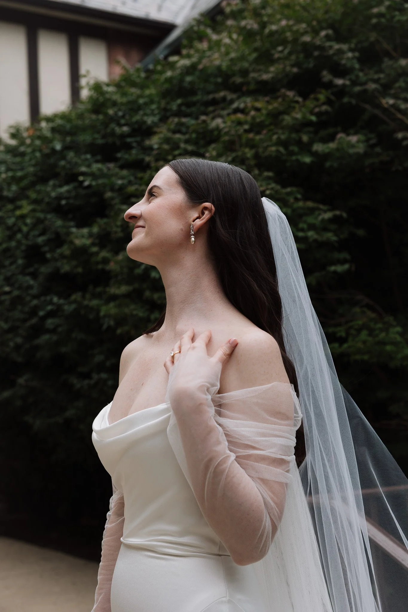 Bride in white wedding dress, with veil and pearl earrings, smiling softly, standing outdoors near a green leafy background.