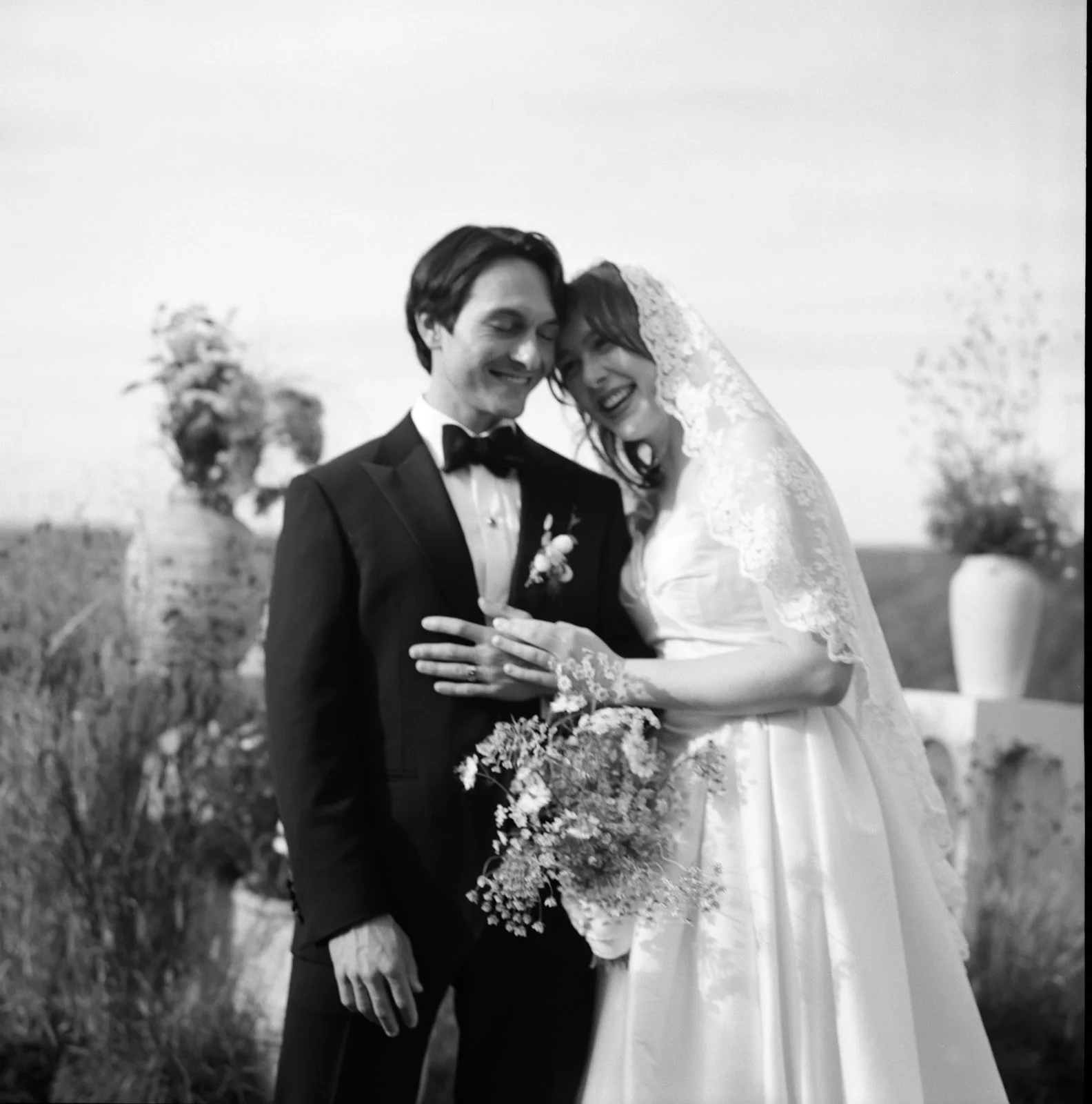 Black and white photo of a bride and groom smiling and holding each other, outdoors, with flowers in the background.